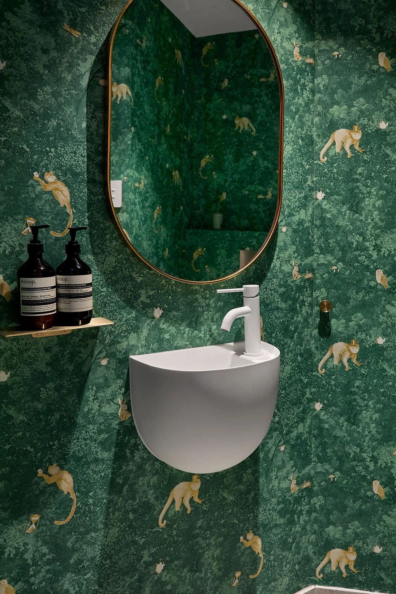 A bathroom with green jungle-themed wallpaper featuring illustrations of monkeys and other animals, a white sink with a modern faucet, a large oval mirror with a thin gold frame, and a small wall-mounted shelf holding two bottles of hand soap.