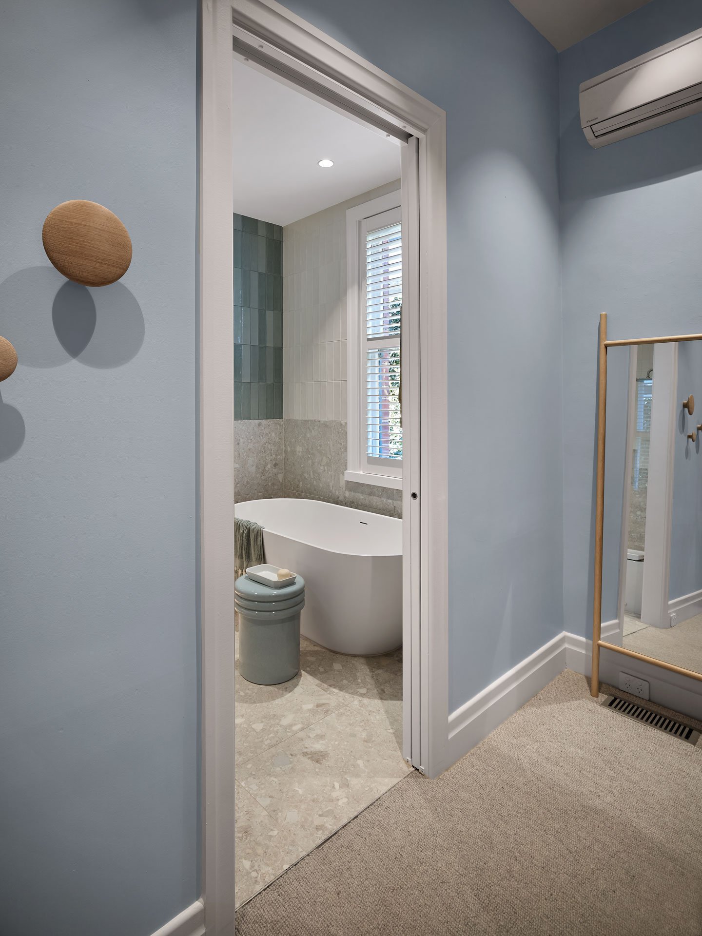 A view of a bathroom with a white freestanding bathtub, a window with shutters, and a greenish-gray tiled wall. The bathroom is accessed through a door from a room with light blue walls and a beige carpet. There is also a small light blue stool or trash can with a white soap dish on top, and a wooden hat or accessory holder mounted on the wall in the adjacent room.