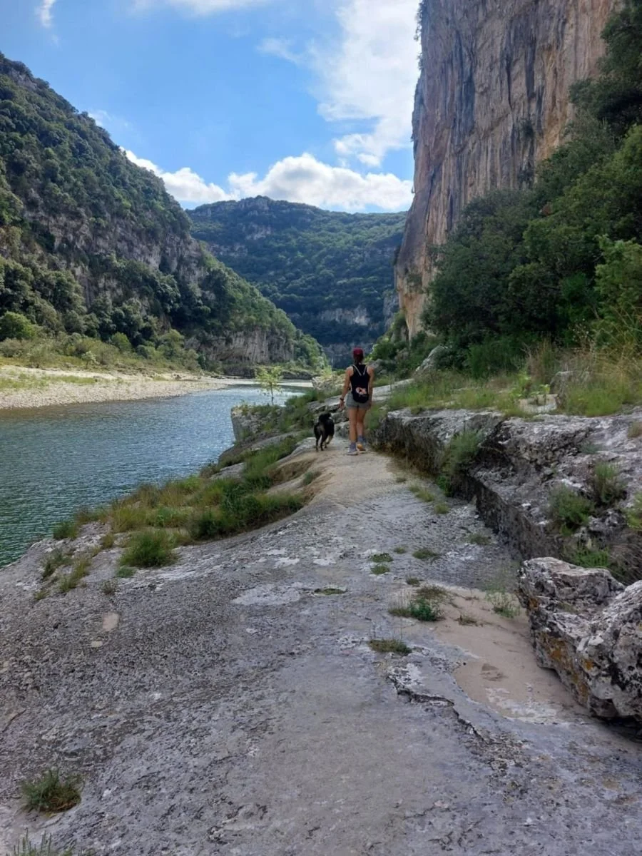 Vrouw wandelt met hond langs een rivier tussen groene bergen tijdens een rustige vakantie, symbool voor ontspanning en verbinding met de natuur.