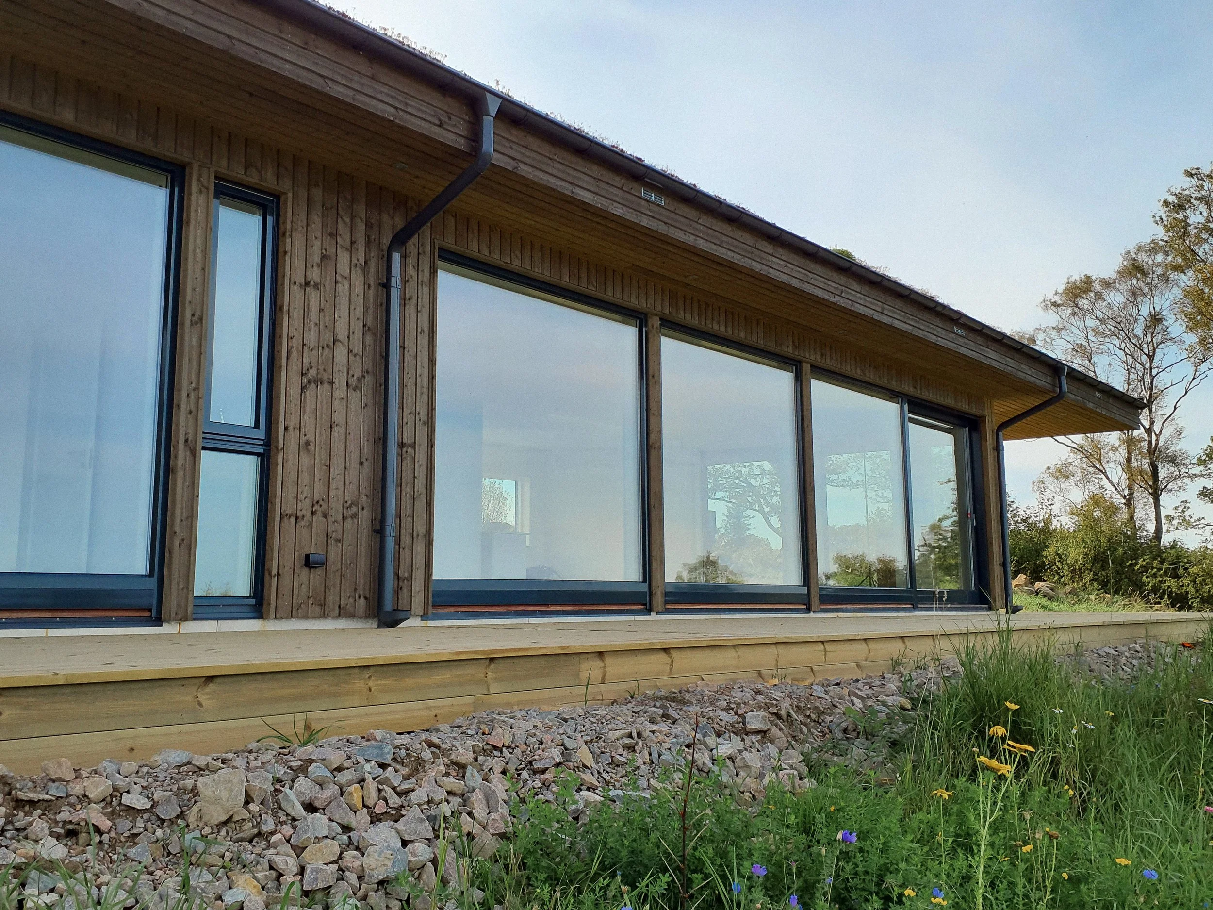 Detta är en modern träbyggnad med stora glasfönster, en träplattd veranda och omgivande grönska och blommor.