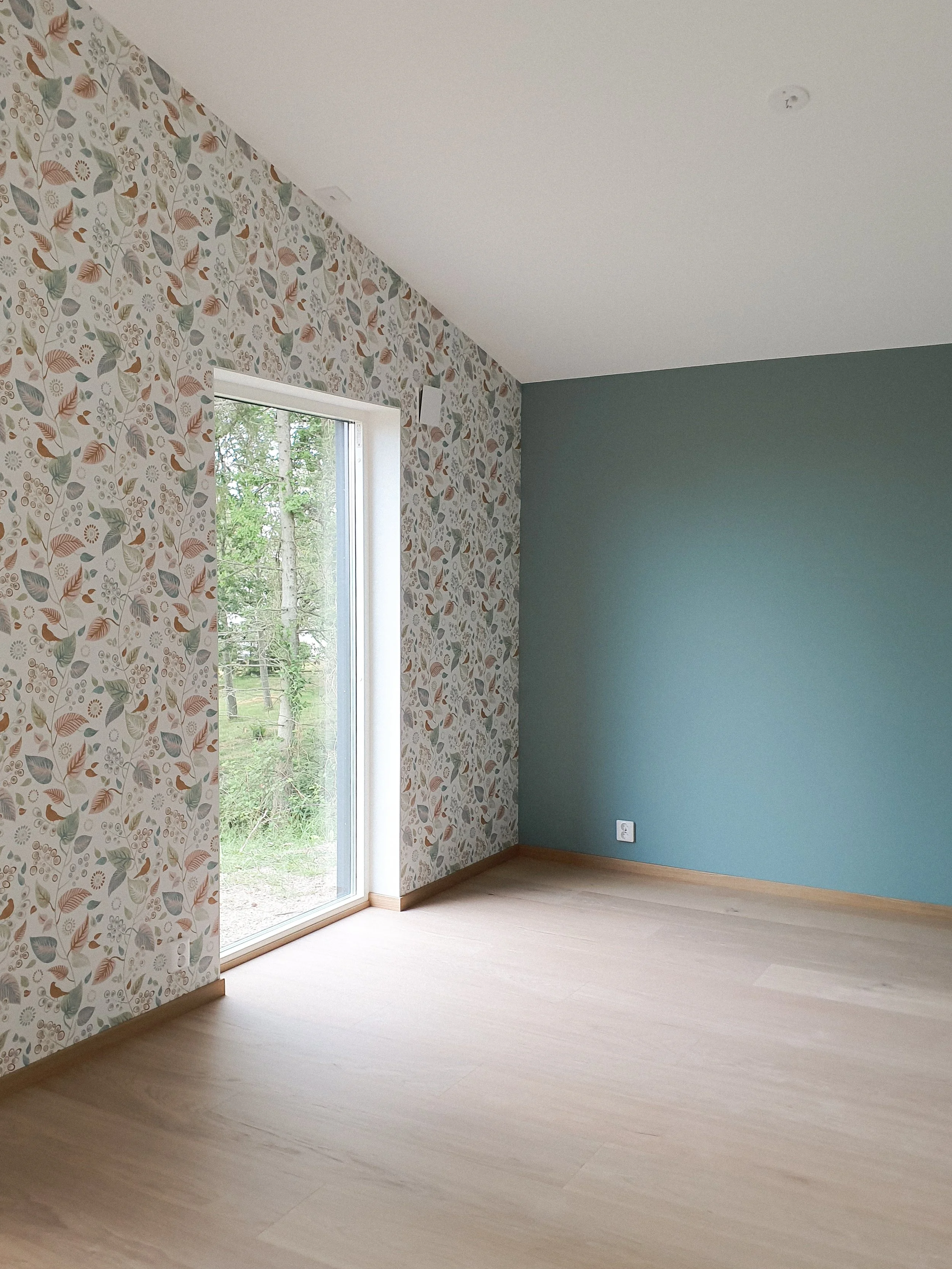 Ett tomt rum med tapetserade väggar, ett stort fönster, trägolv och eluttag.