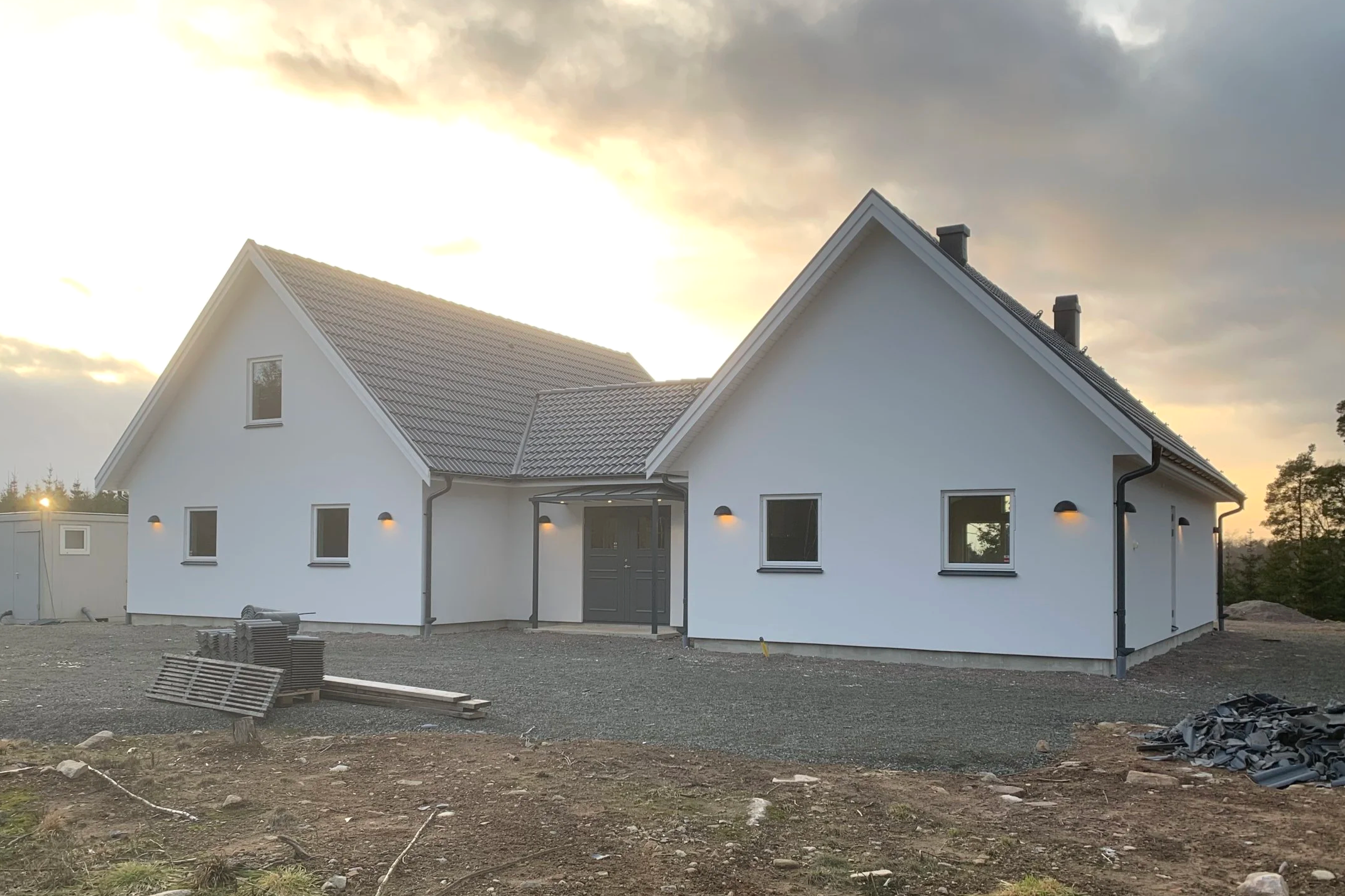 Färdigställt vitt hus med mörk takpanna och utomhusbelysning, under uppbyggnad, på en tomt.