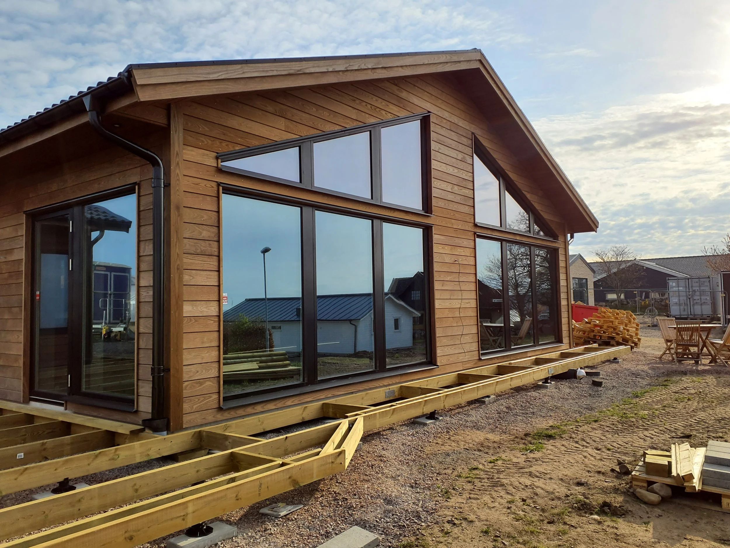 Nybyggd trähus med stora fönster och delar av trädäck under konstruktion, byggmaterial och utemöbler runt omkring.