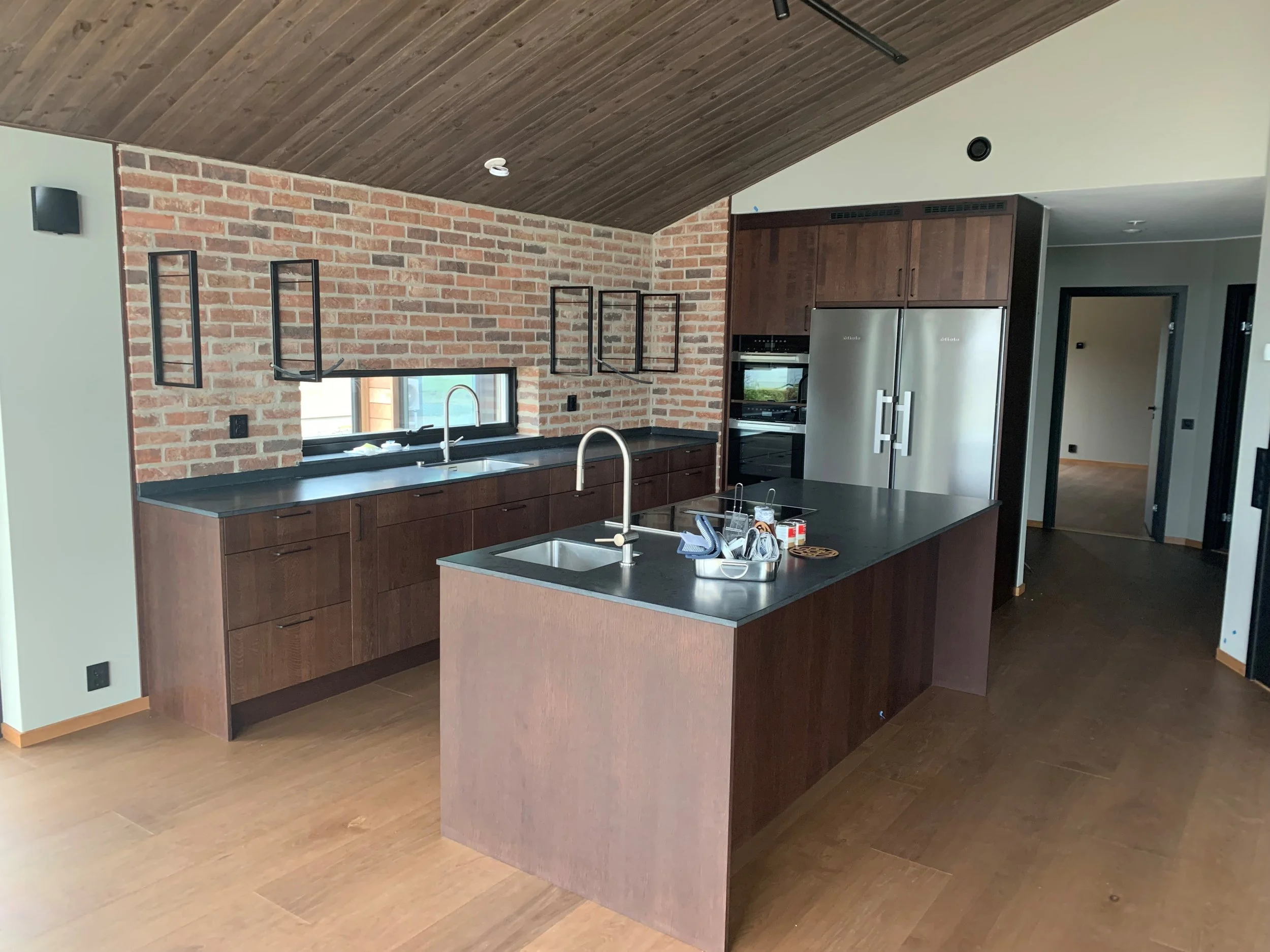 Modernt kök med mörka trämöbler, svart bänkskiva, öppna skåp och ett brickbord, bricker i fönsteren och en metallintegrerade hylla över diskbänken. Brickbordet har olika köksredskap och förbrukningsvaror.