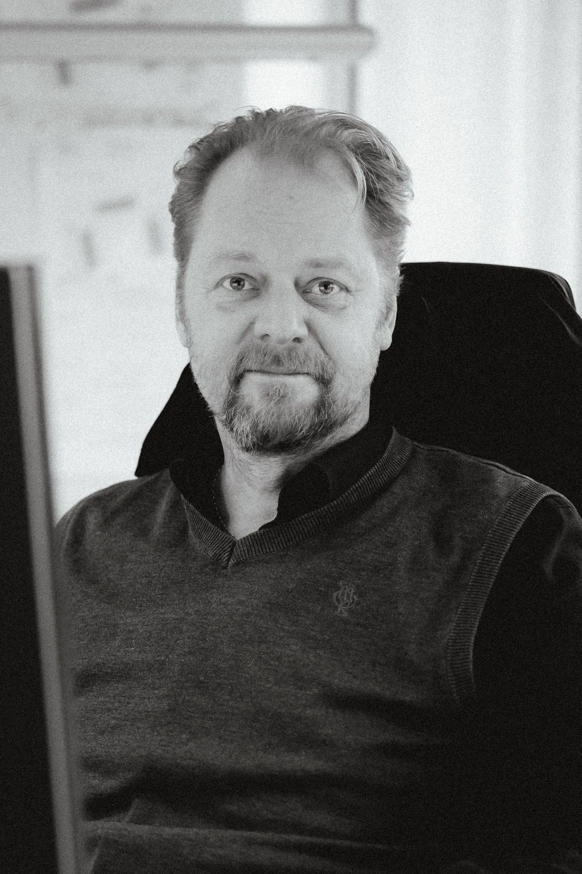 Svartvitt foto av en man med skägg och ljus hår, sittande i en kontorsstol, klädd i en mörk tröja och svart skjorta.