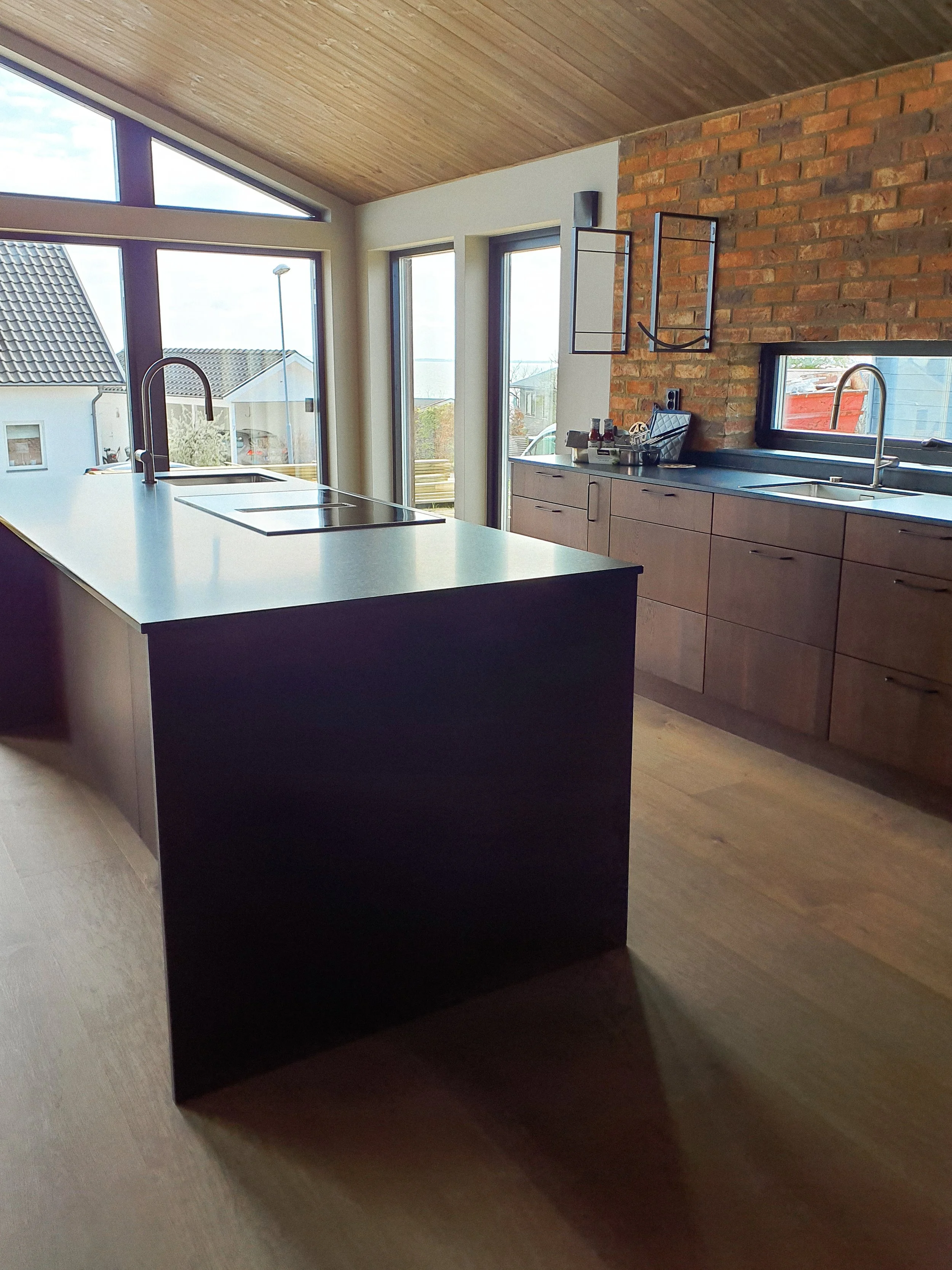 Moderne kök med trästommar, stora fönster och en köksö i mörk färg.