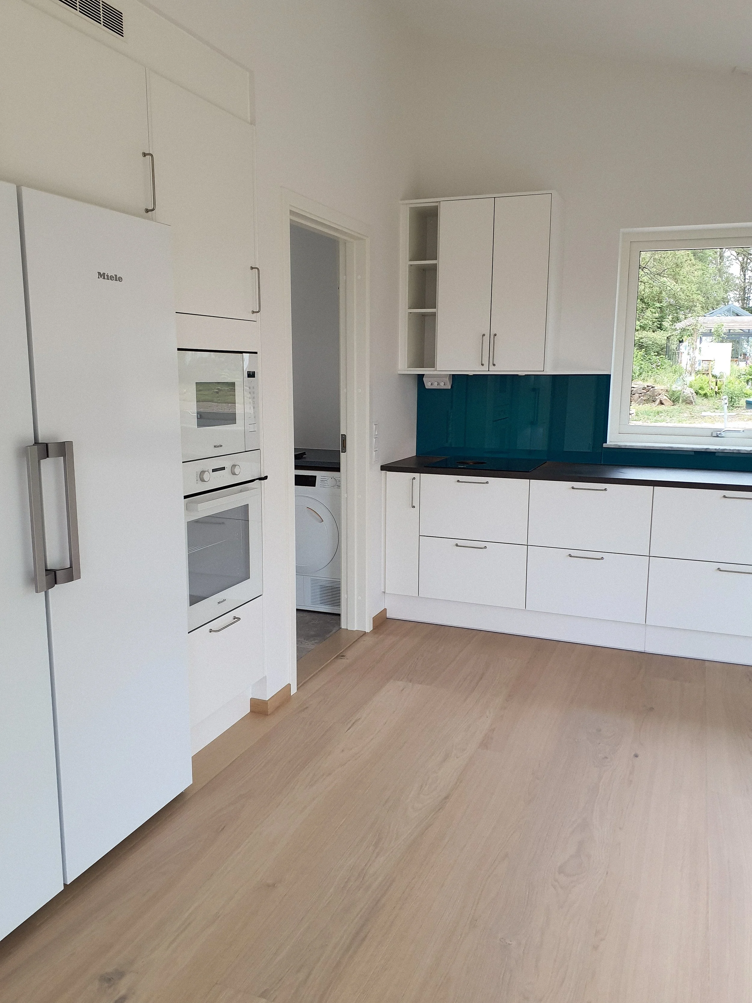 En modern köksinteriör med vita skåp, en svart bänkskiva och ett stort fönster som visar en trädgård utanför.