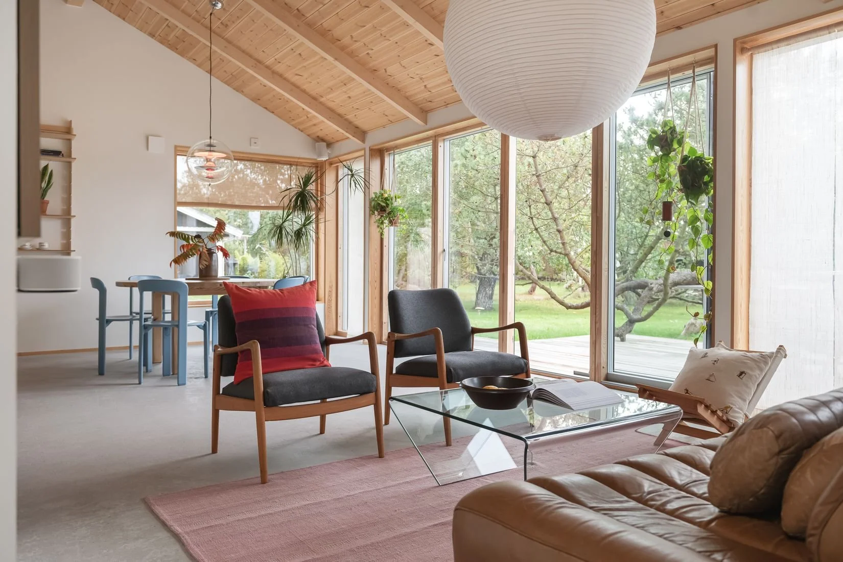 Ljust vardagsrum med stora fönster och utsikt över träd och gräsmatta, möbler i trä och tyg, samt tak av träpanel.