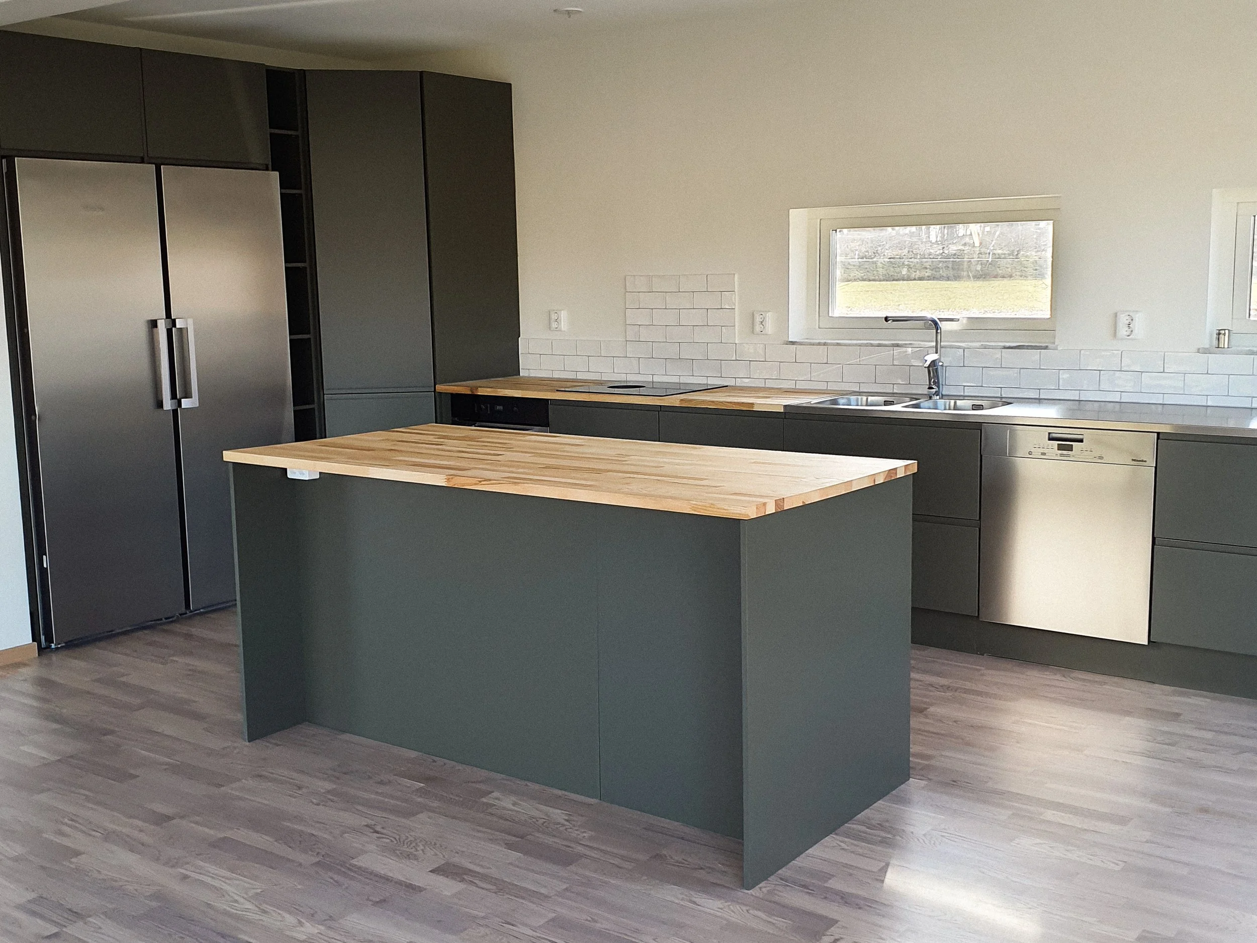 Modernt kök med grå skåp, vit kakelvägg, vinylgolv, köksö med träbänkskiva, fönster över vasken och diskmaskin.