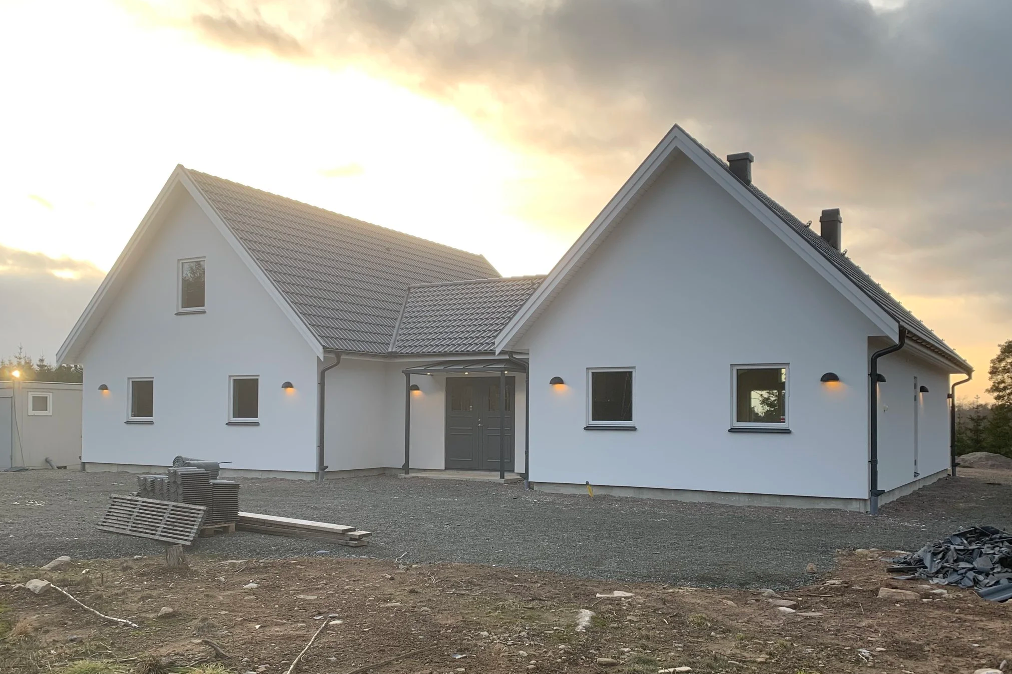 En vit nybyggd villa med svart tak, tre fönster på framsidan, utomhusbelysning, pågående utomhusarbete med byggmaterial och markarbete