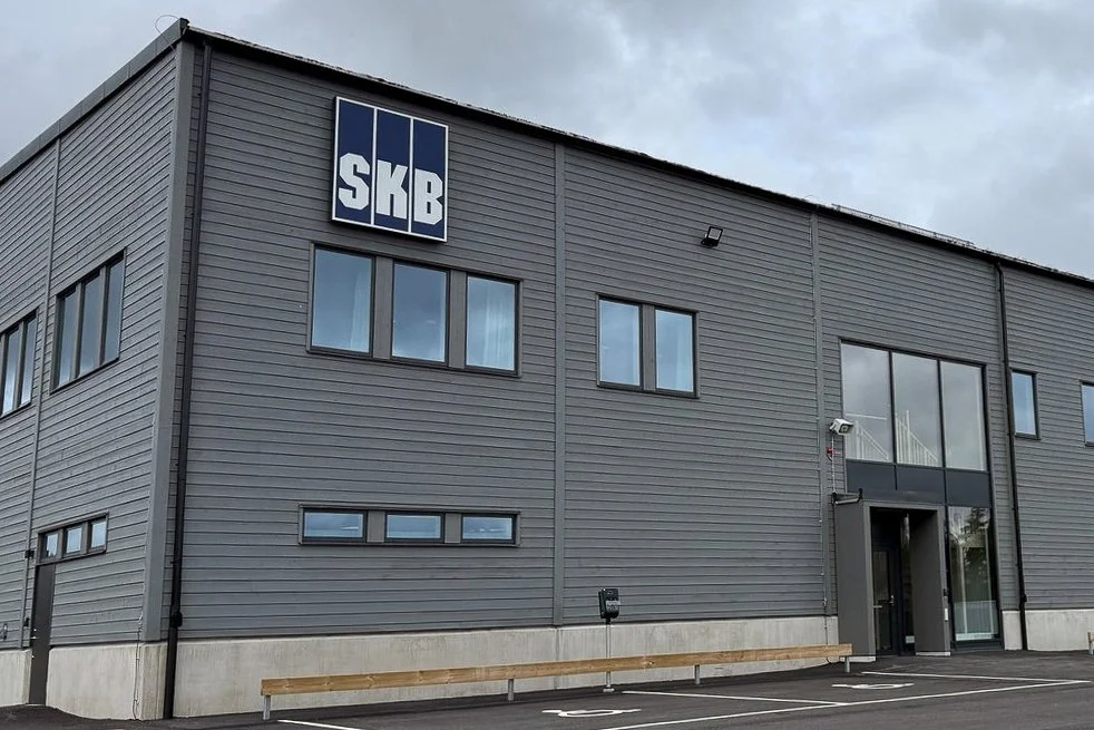 En modern, grå byggnad med SKB-logotyp nära toppen, flera fönster och en entré med glasdörr. Ovanför entrén finns ett kamerahus. Skyar i bakgrunden.