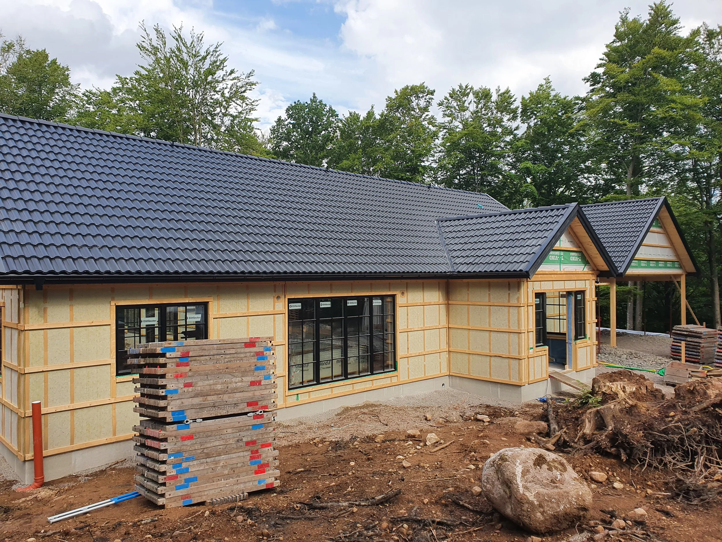 Kuperat hus under byggnation med svart tak, fönster och träskyddsfolie på väggarna, omgiven av byggmaterial och jord.