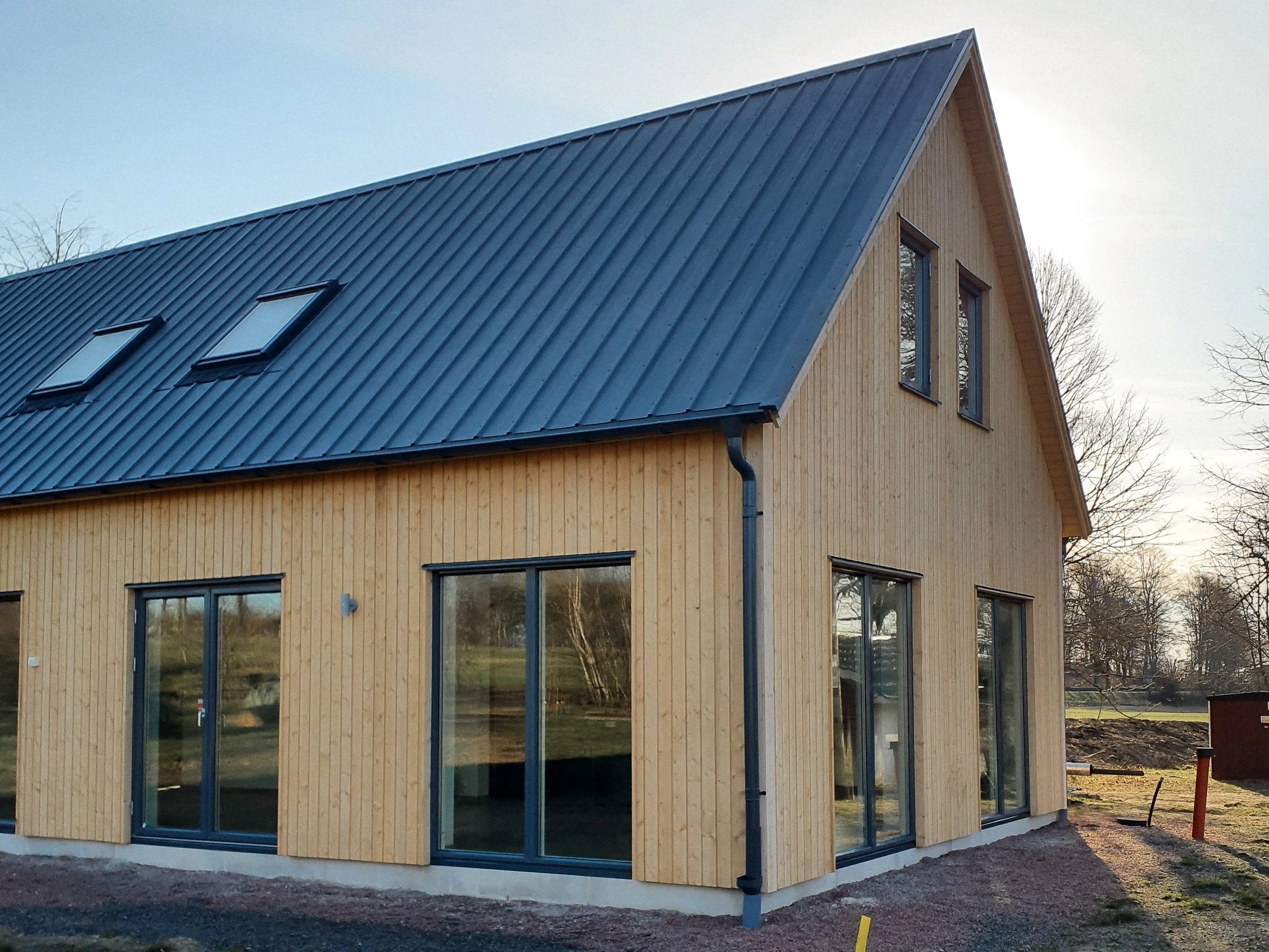 En modern, tvåvåningshus under byggnation med träfasad och blå metalltak, stora fönster och tre takfönster.