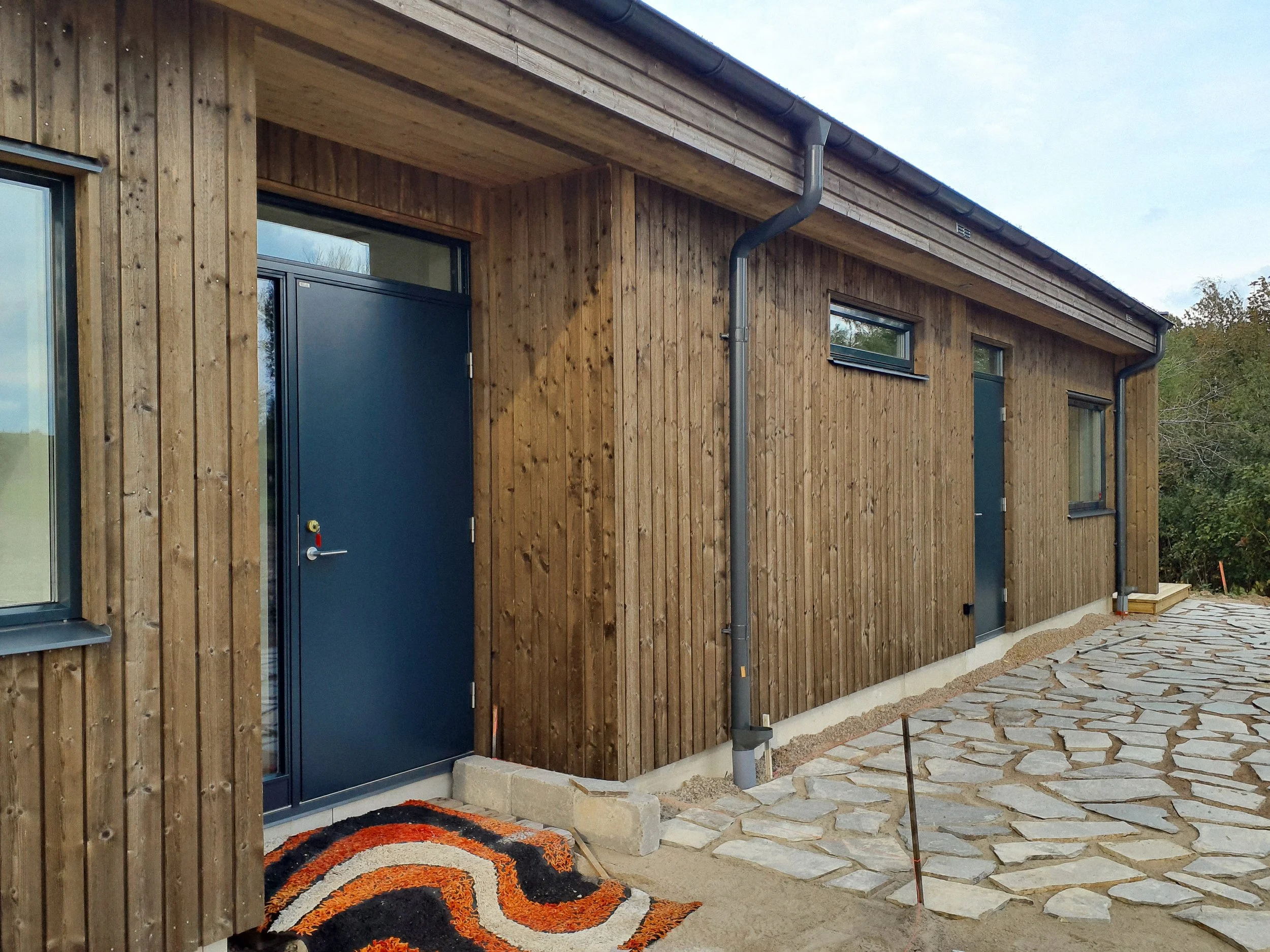 Exteriör av ett nybyggt trähus med mörkblå dörrar och små fönster, samt en stenlagd gångväg framför.