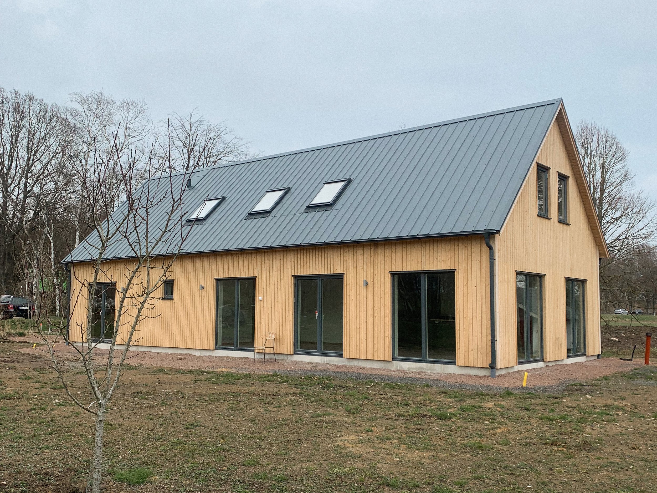 Ett nybyggt hus med träfasad och svart metalltak, omgiven av ett öppet jord-och-gräsområde och några träd, under en molnig himmel.