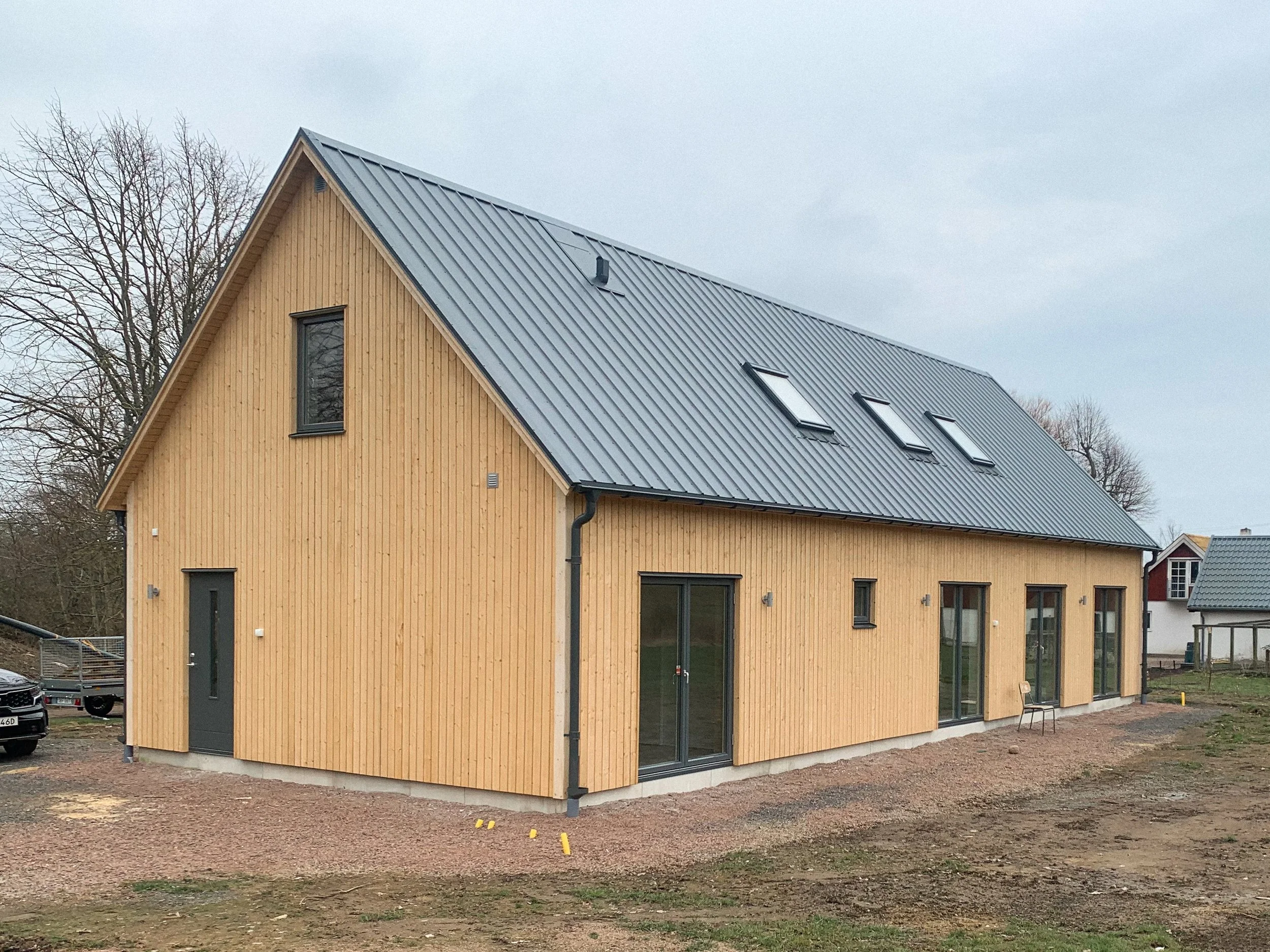 En nybyggd trävilla med ljus träfasad och metalltak, omgiven av en tom tomt.