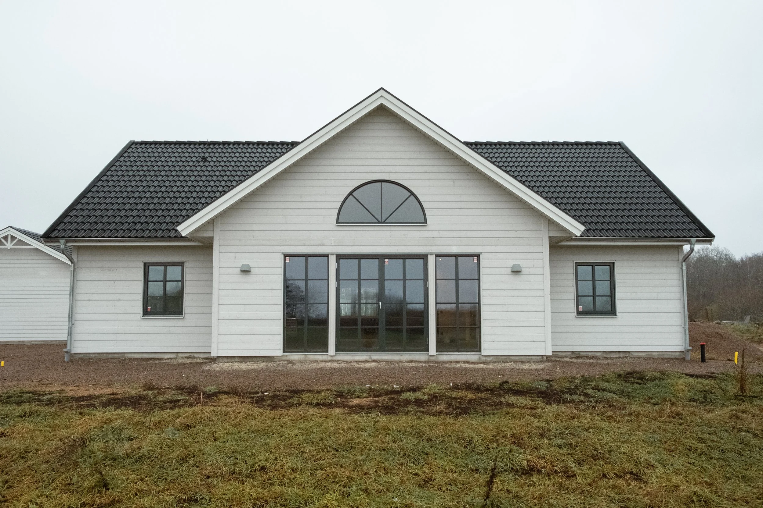 Nyanbyggt vitt hus med svarta tak, stora fönster och dörrar, byggd på en tomt med gräs och jord, omgiven av ett landskap med träd i bakgrunden.