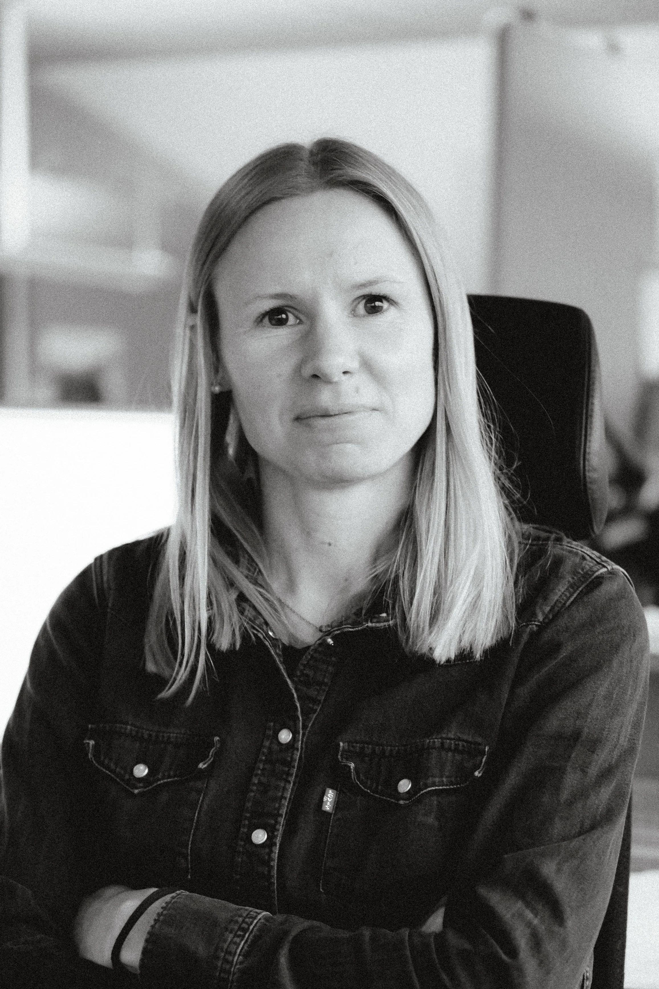 Svartvitt porträtt av en kvinna med axellångt blont hår, sittande vid ett skrivbord i ett kontor, med armarna korsade.