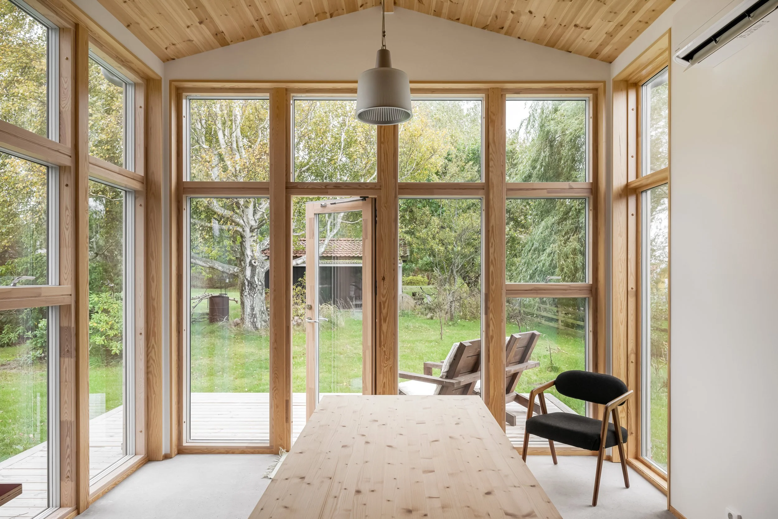 Ett inomhusutrymme med stora fönster och trädgård utanför, ett bord och en svart och trästol.
