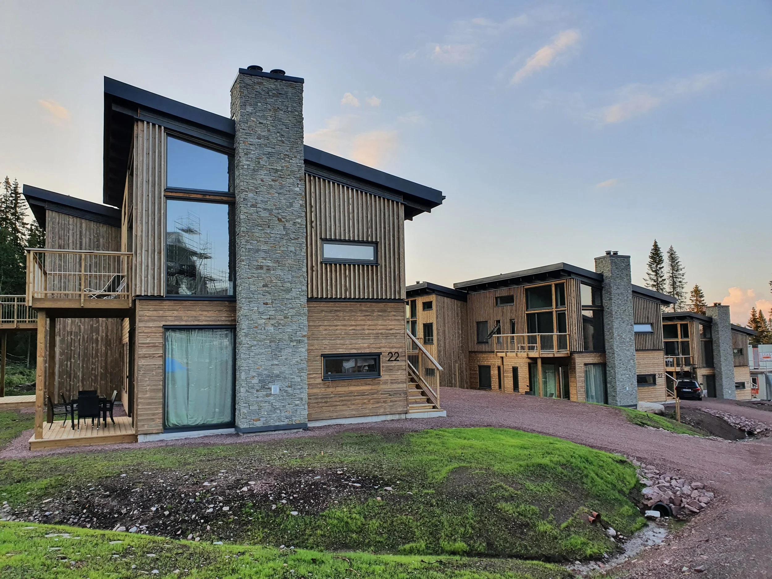 Nya moderna flerfamiljshus med trä och stenfasad, stora fönster, små balkonger och garage, omgivna av grön mark och träd i bakgrunden.