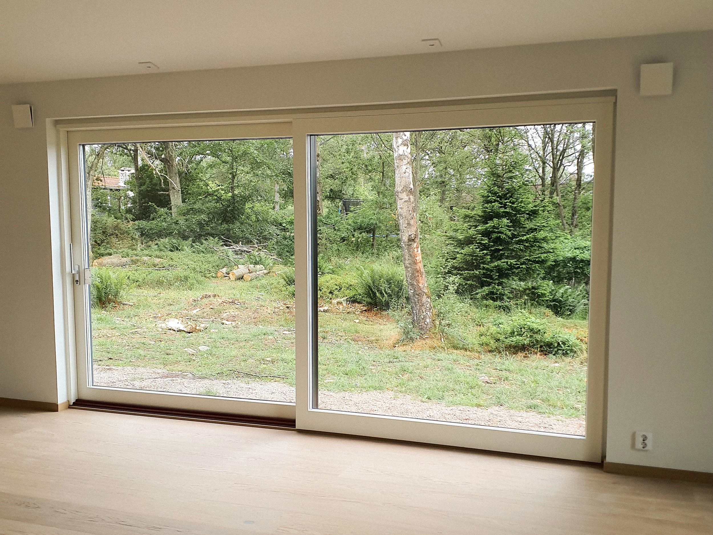 Ett tomt rum med stora glasdörrar som leder till en trädgård med gröna träd och gräs.