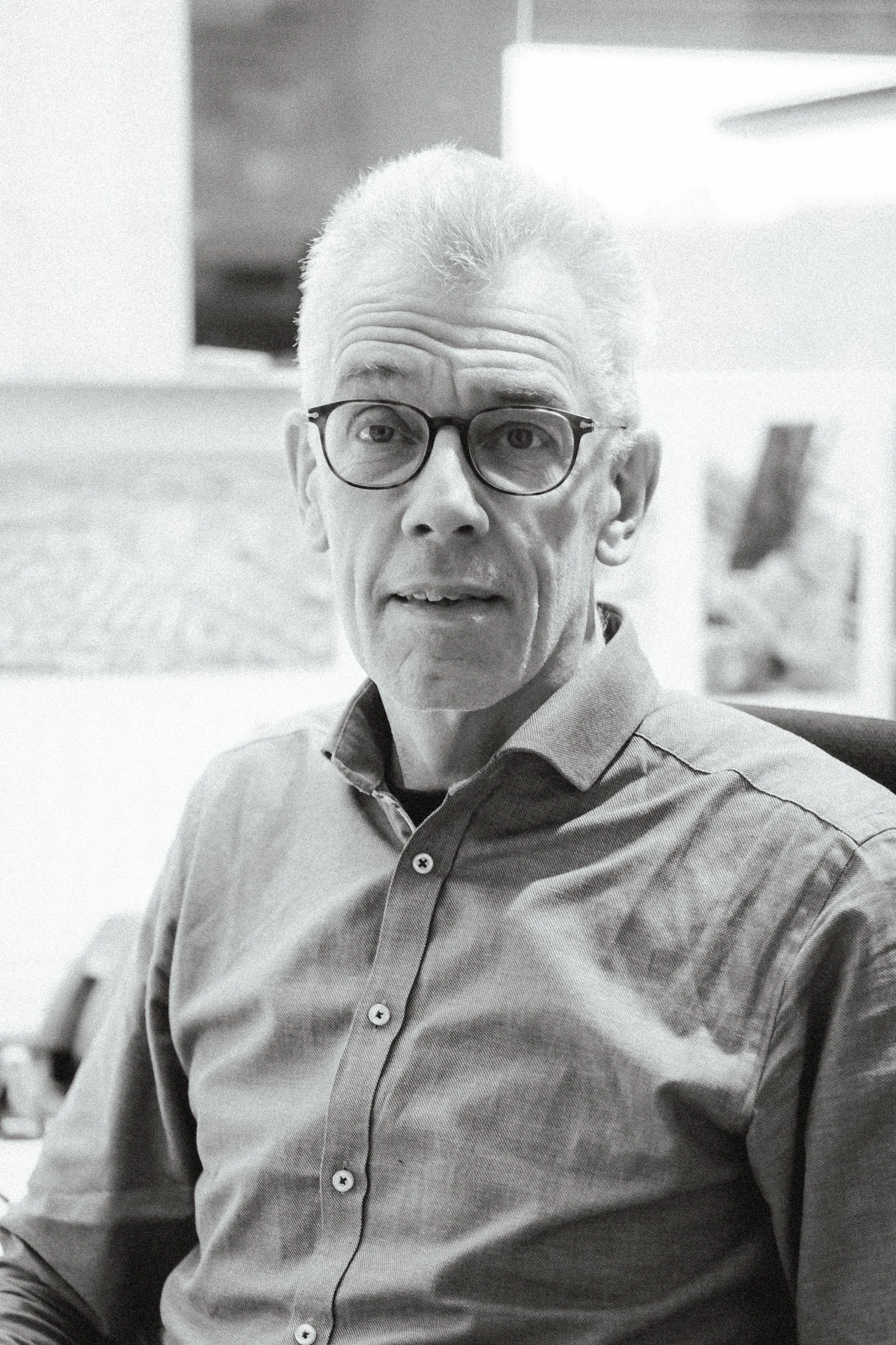 En äldre man med glasögon, grått hår, sittandes i ett kontorsmiljö.