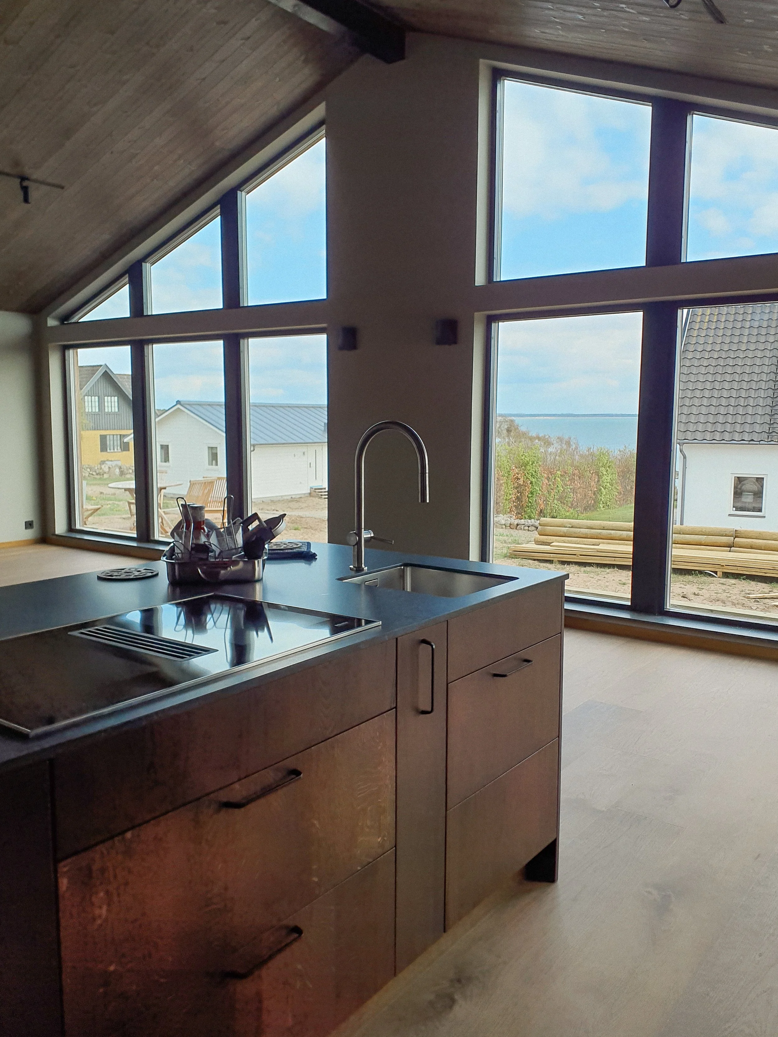 Kök med stort fönster som visar en trädgård och hav i bakgrunden, inomhus med trägolv och modern köksö.