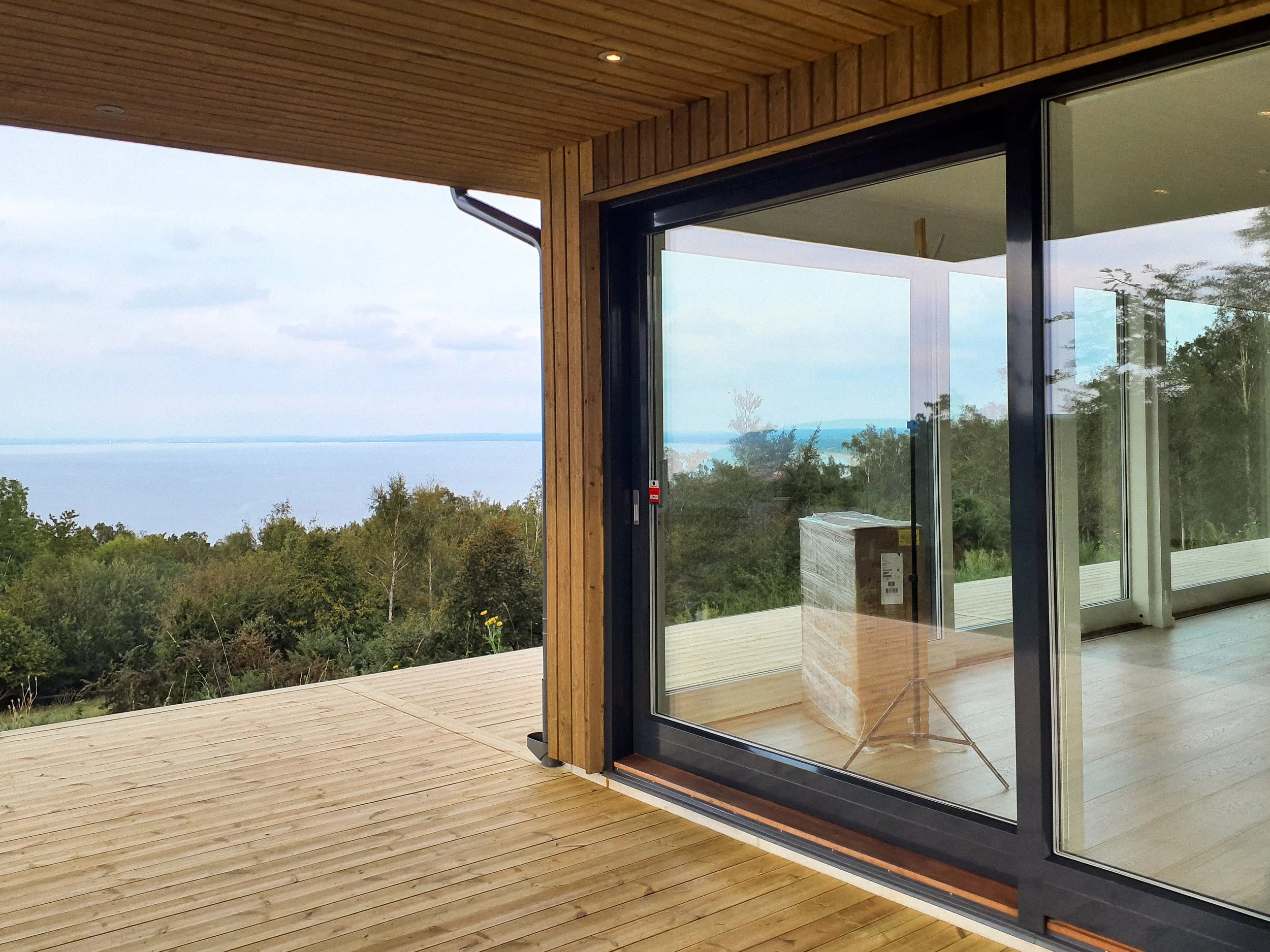 En modern trävåning med stor glasdörr och utsikt över skog och vatten.