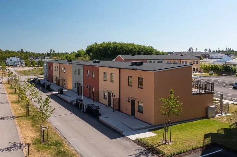 Flerfamiljshus i modern stil med flera sommarstugor i bakgrunden, omgivna av små träd och tillgång till p-platser