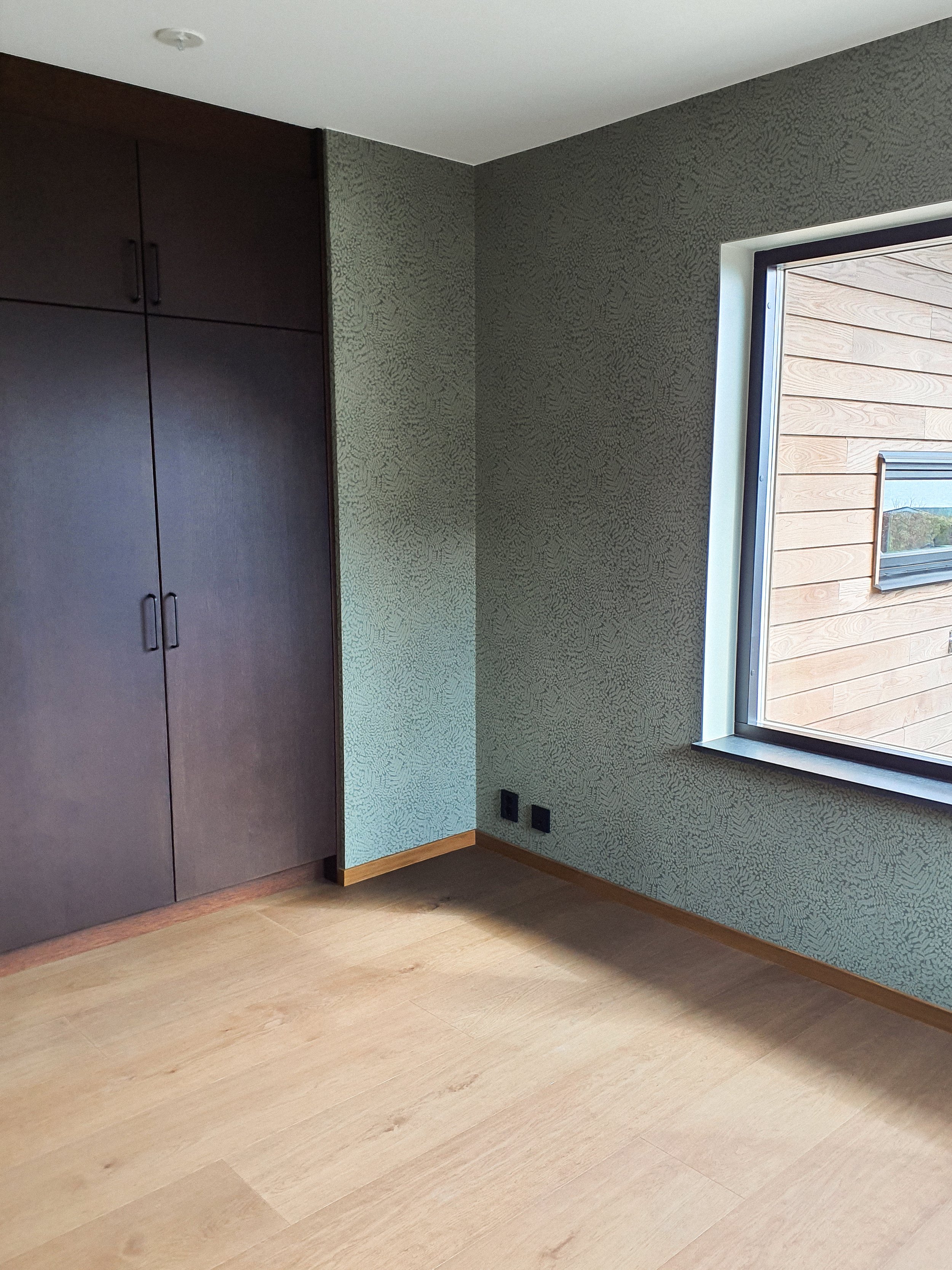 Ett tomt rum med trägolv, en stor fönstervägg, ett mörkt garderobsskafferi, väggar med mönstrad tapet och ett eluttag nära golvet.