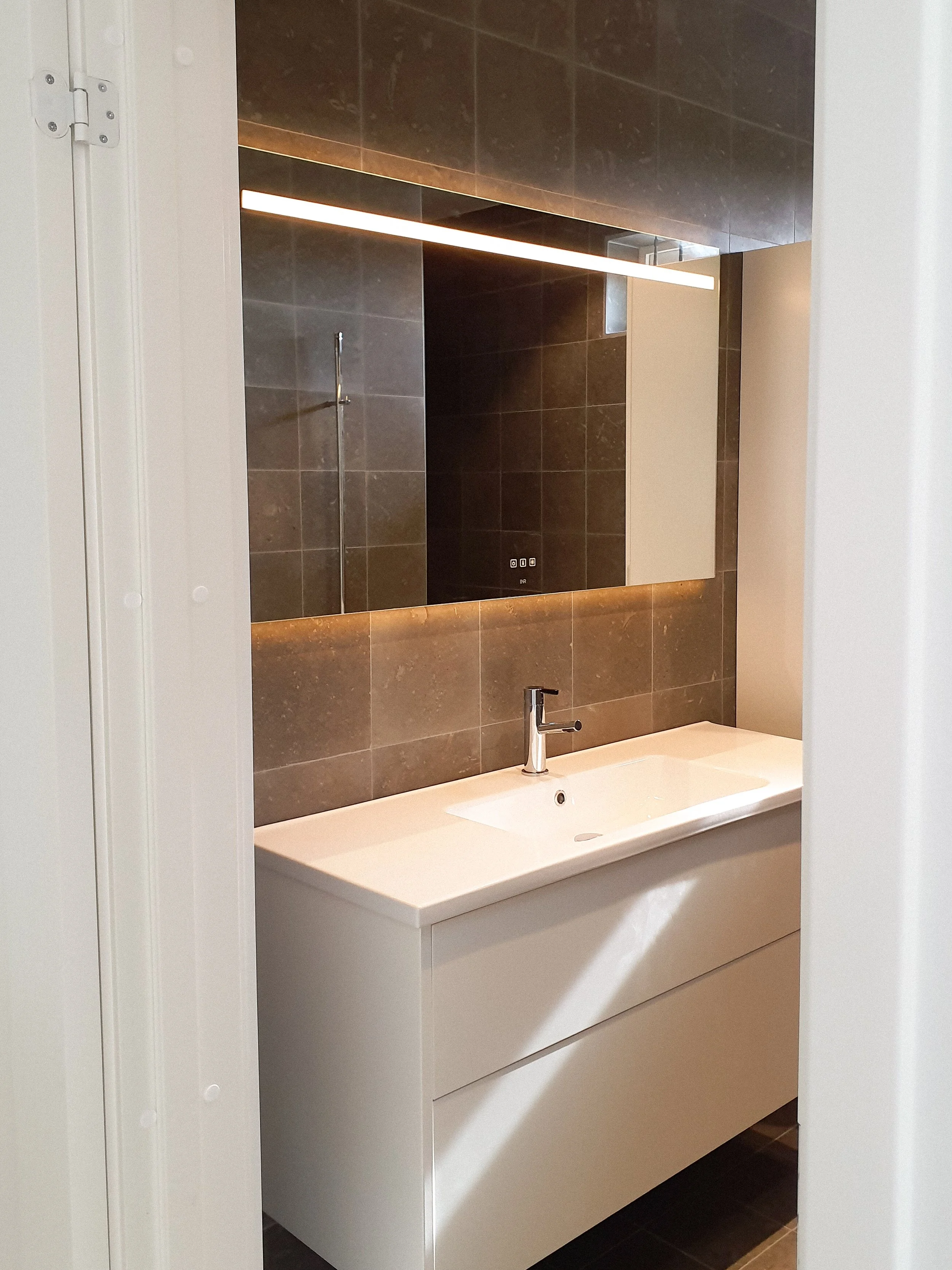 En modern badrumsspegel med inbyggd belysning ovanför tvättbänk med handfat.