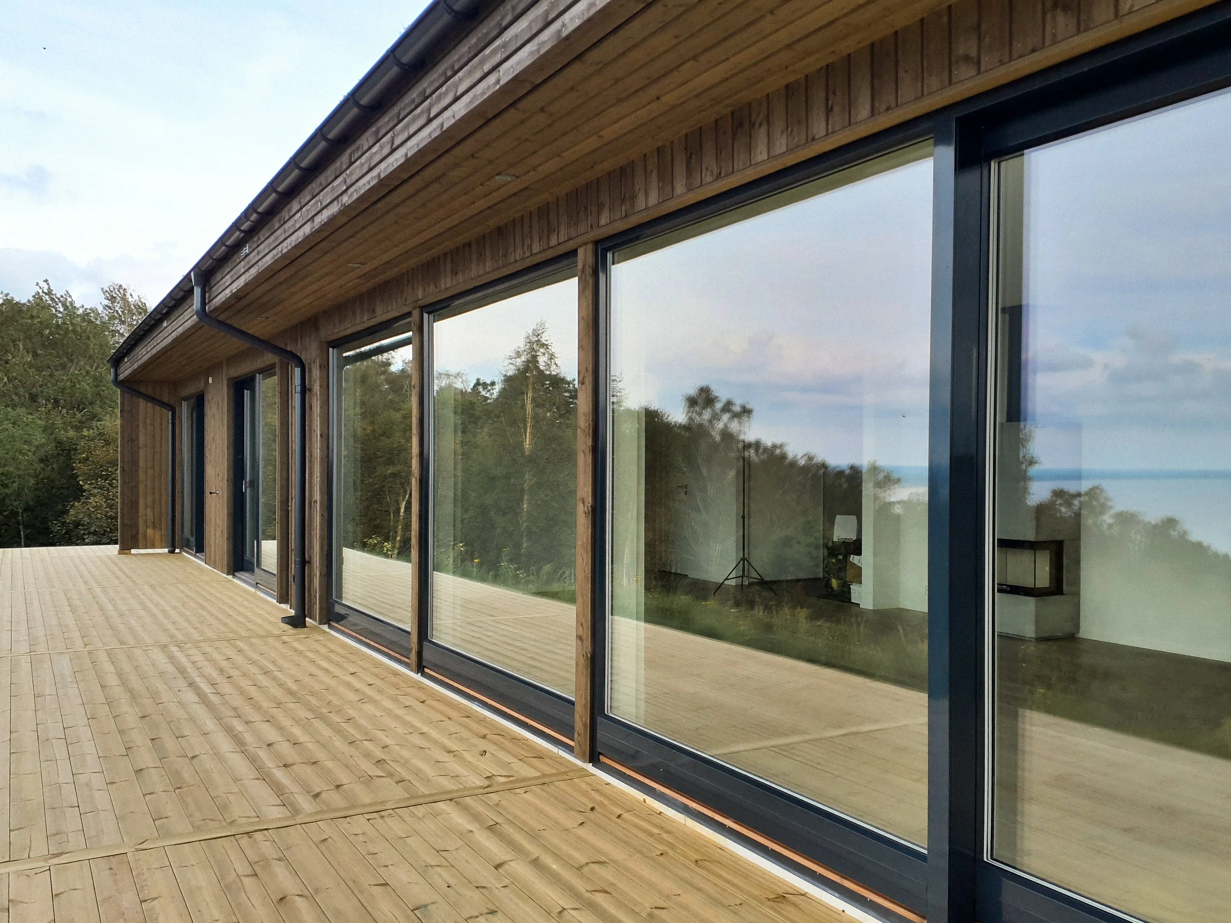 En träaltan framför ett modernt hus med stora glasfönster och en trädgård med träd i bakgrunden.