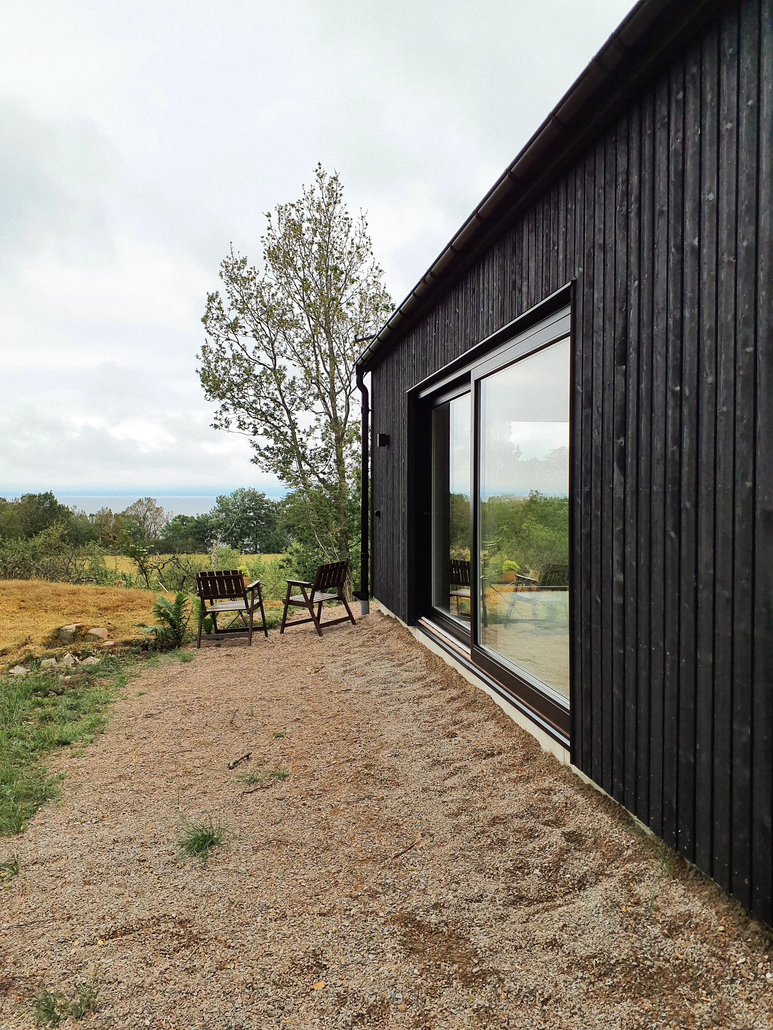 Utsikt över ett modernt svart hus med stora fönster, omgiven av en terrass med två trästolar och ett träd i bakgrunden, under en molnig himmel.