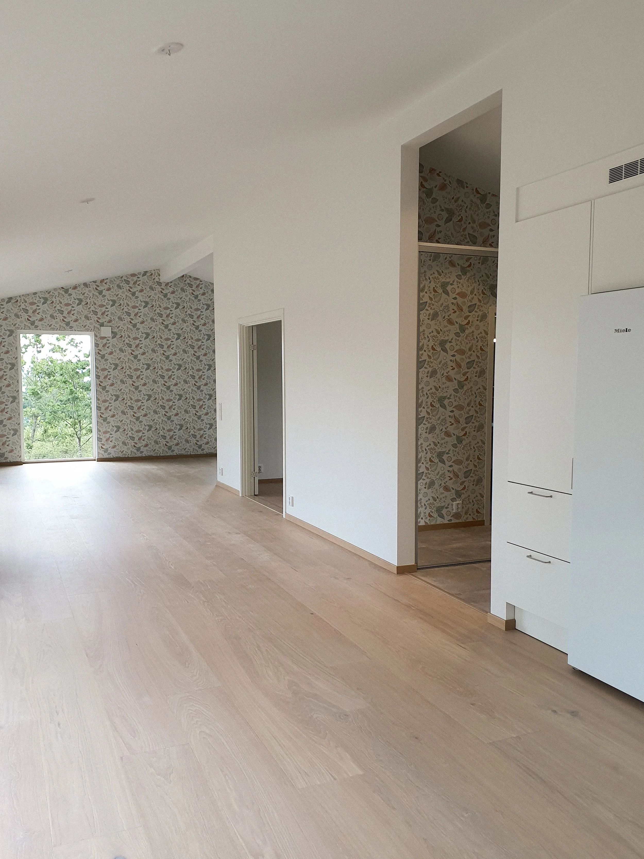 Ljust inredd vardagsrum med ljus trägolv och väggar, ett fönster med utsikt över grön natur samt en innervägg med tapet med blomster- och bladmotiv.