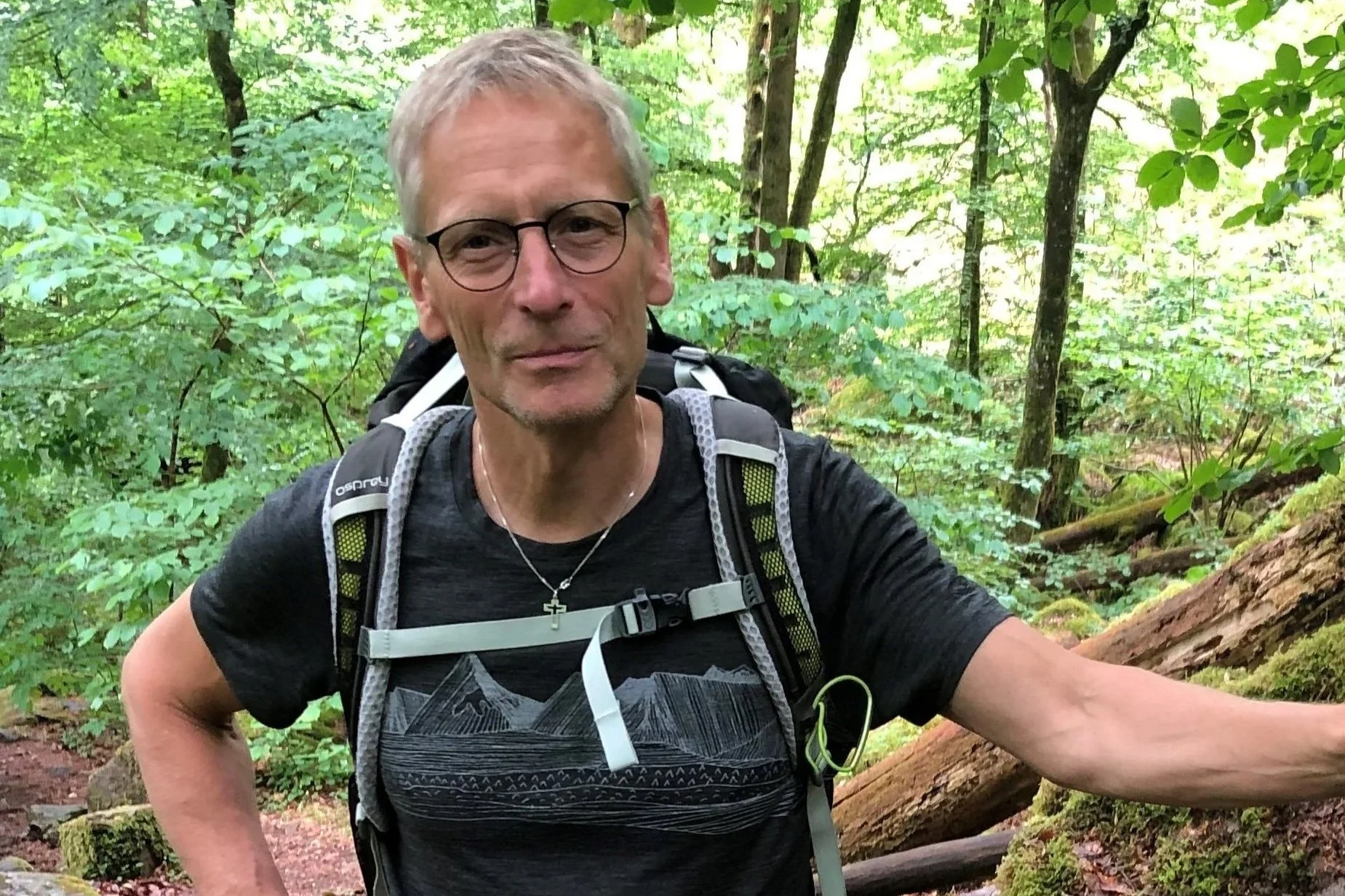 En man med grått hår, glasögon, svart T-shirt och ryggsäck står i en grön skog.