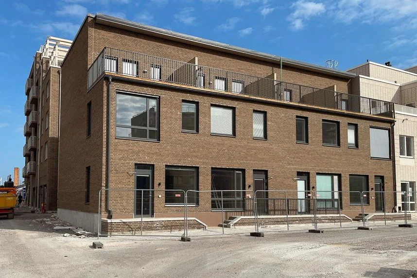En nybyggd flerfamiljsfastighet med brunt tegel, stora fönster och en takterrass med räcken. Pågår byggnadsarbete framför byggnaden.