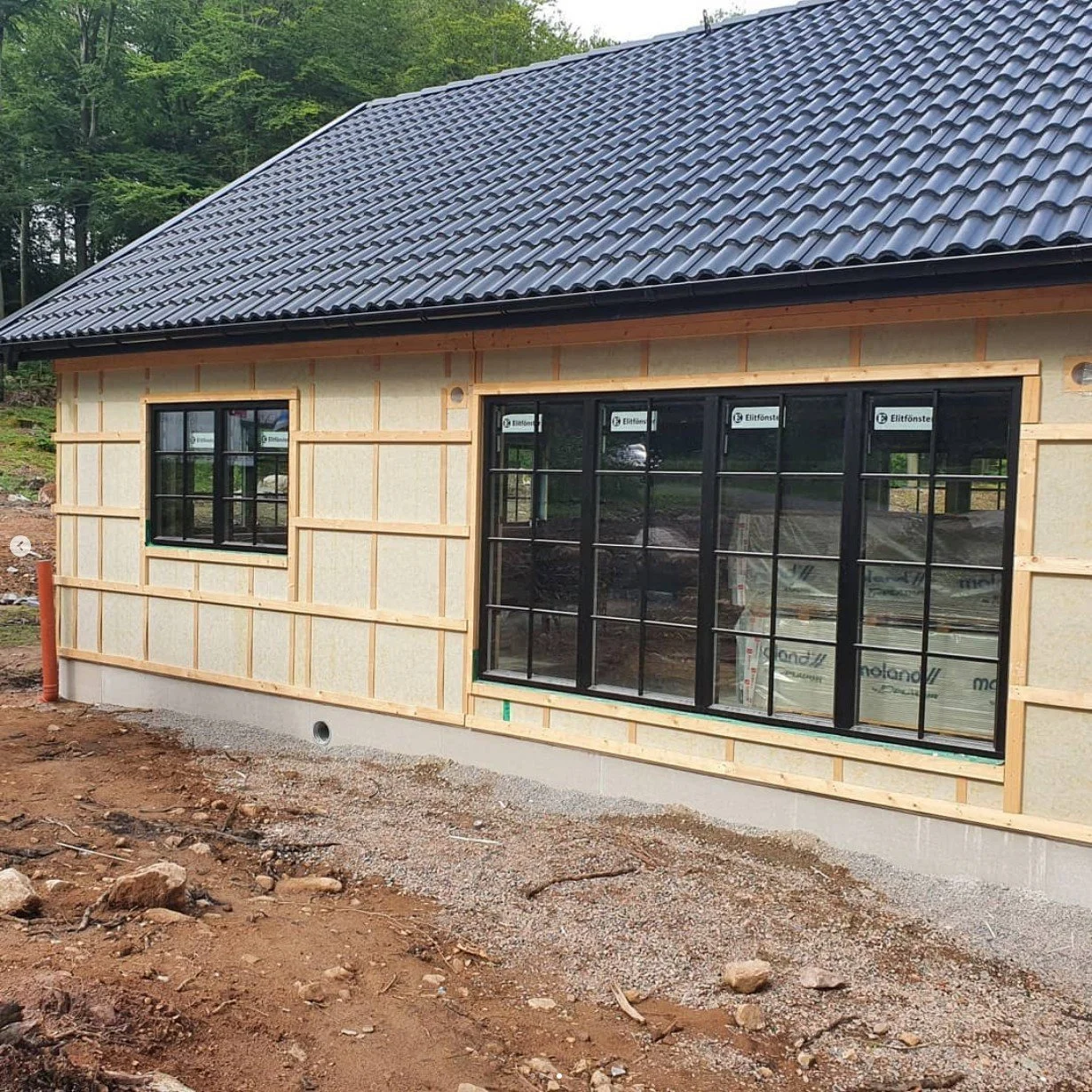 Byggnation av ett hus med stora fönster, träskivor på yggväggarna och ett tegeltak, omgivet av grön skog.