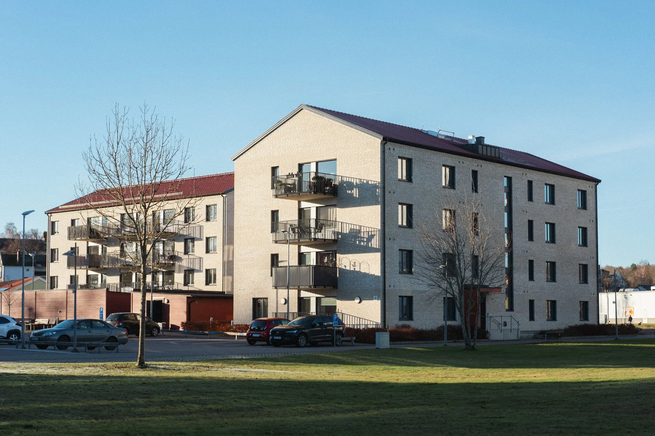 Två moderna flerfamiljshus med ljus tegel, tre våningar, med balkonger, omgivna av park och parkeringsplatser, under en klar blå himmel.