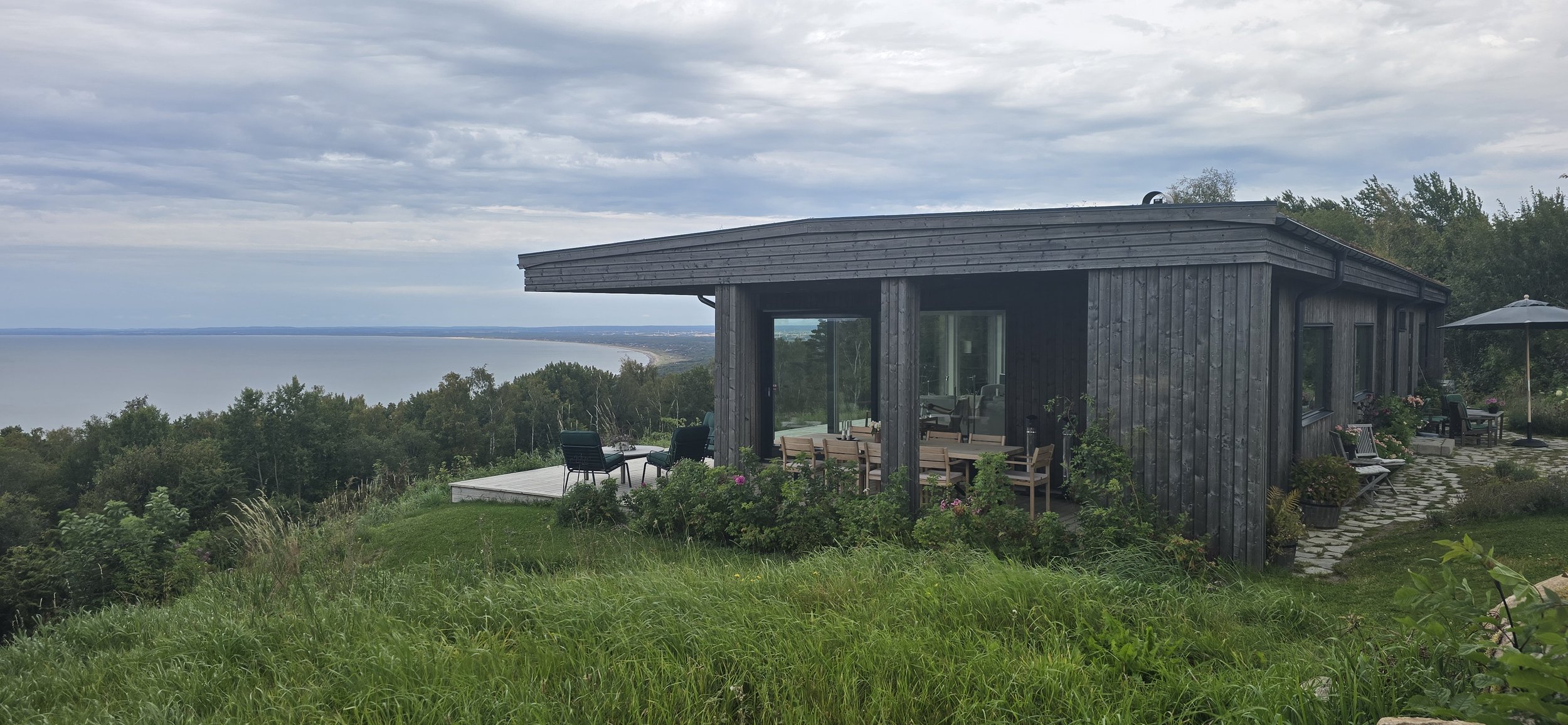 En modern svart trähus på en grön kulle med utsikt över ett stort vatten, utomhusmöbler, parasoll och växter.