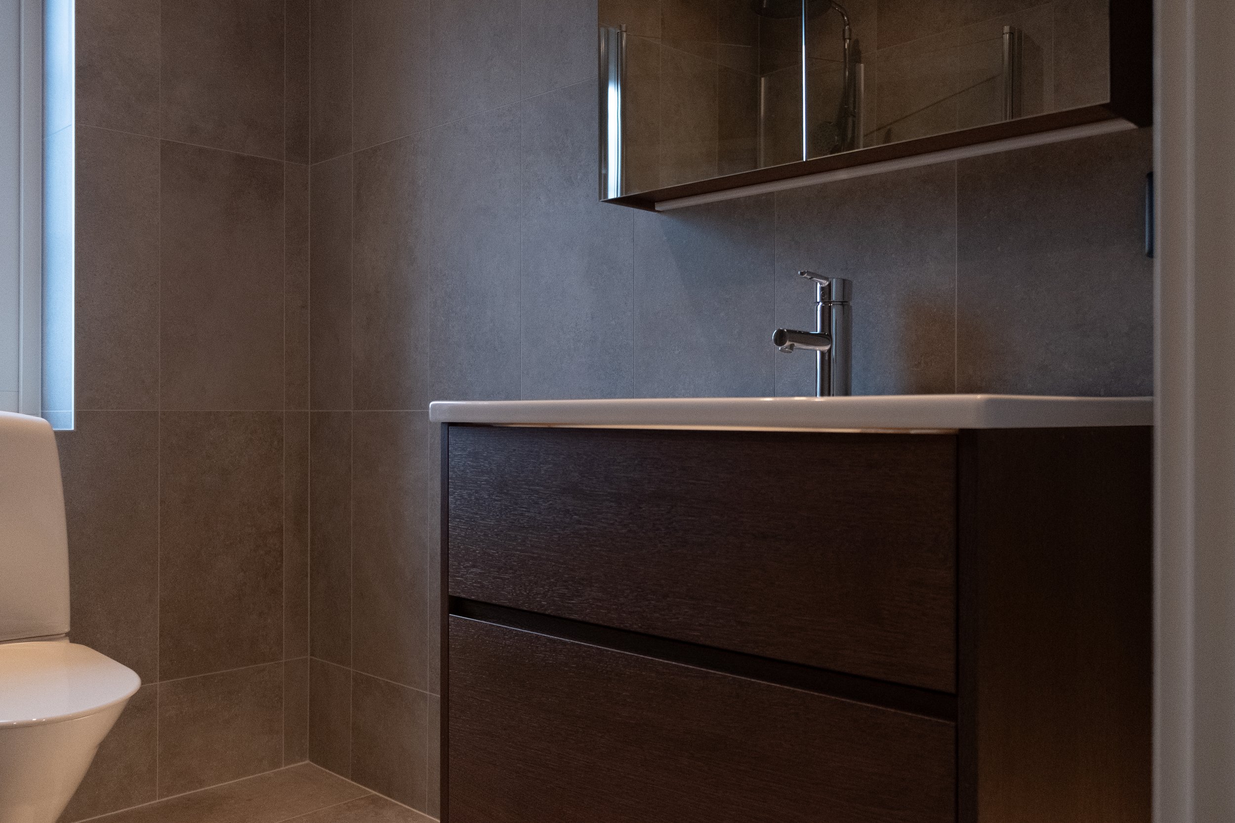 Badrum med mörkbrun skåp, vit handfat, spegel och del av toalett.