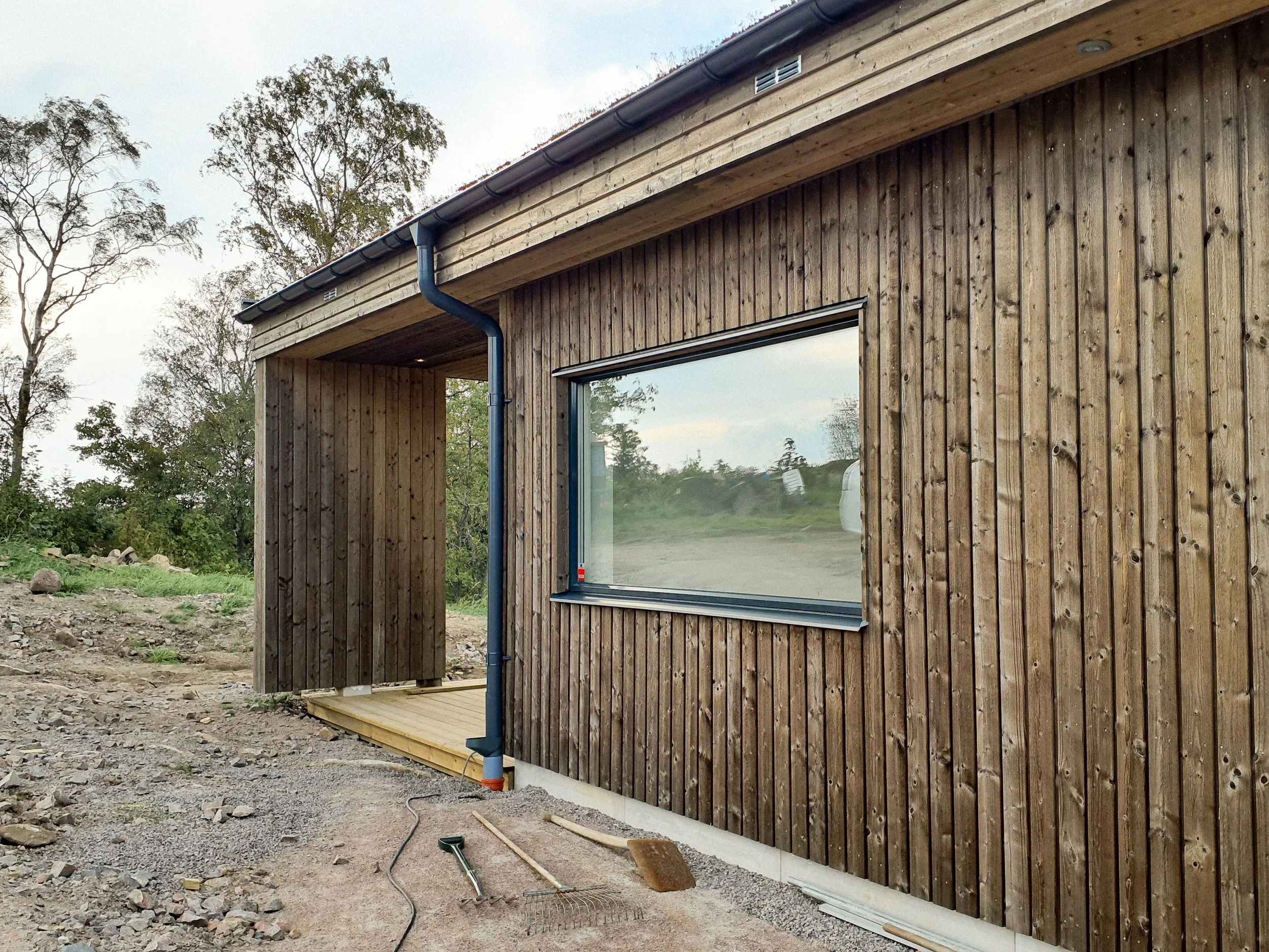 En trävillabyggnad med ett stort fönster och hölje av träpaneler, byggnadens utsida är under konstruktion med arbetsverktyg och material framför.