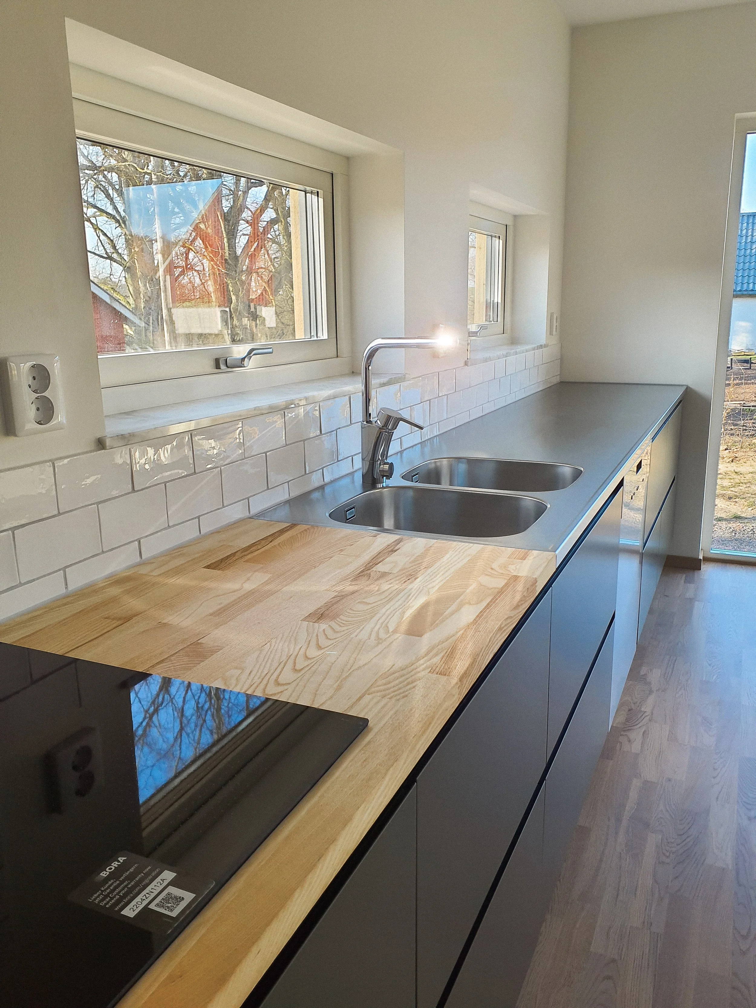 Köksbänk med dubbelho i rostfritt stål, nybyggt kök med vita kakelväggar och två fönster, en med utsikt mot ett vinterträd och ett annat mot en trädgård. Det finns en svart spishäll på längre sidan av bänken.