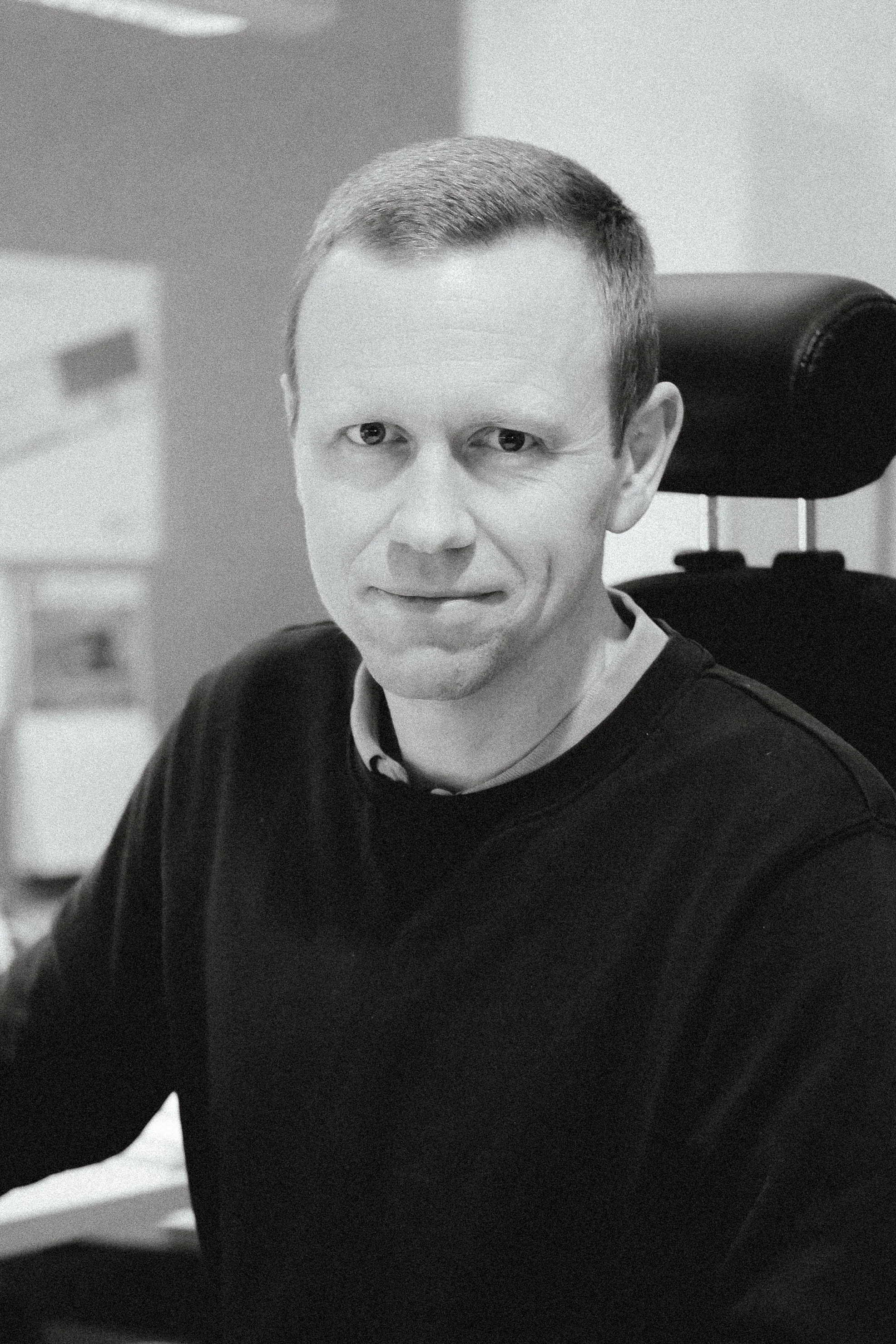 Svartvit porträttbild av man som sitter i en kontorsstol, klädd i svart tröja, med kort hår och leende.