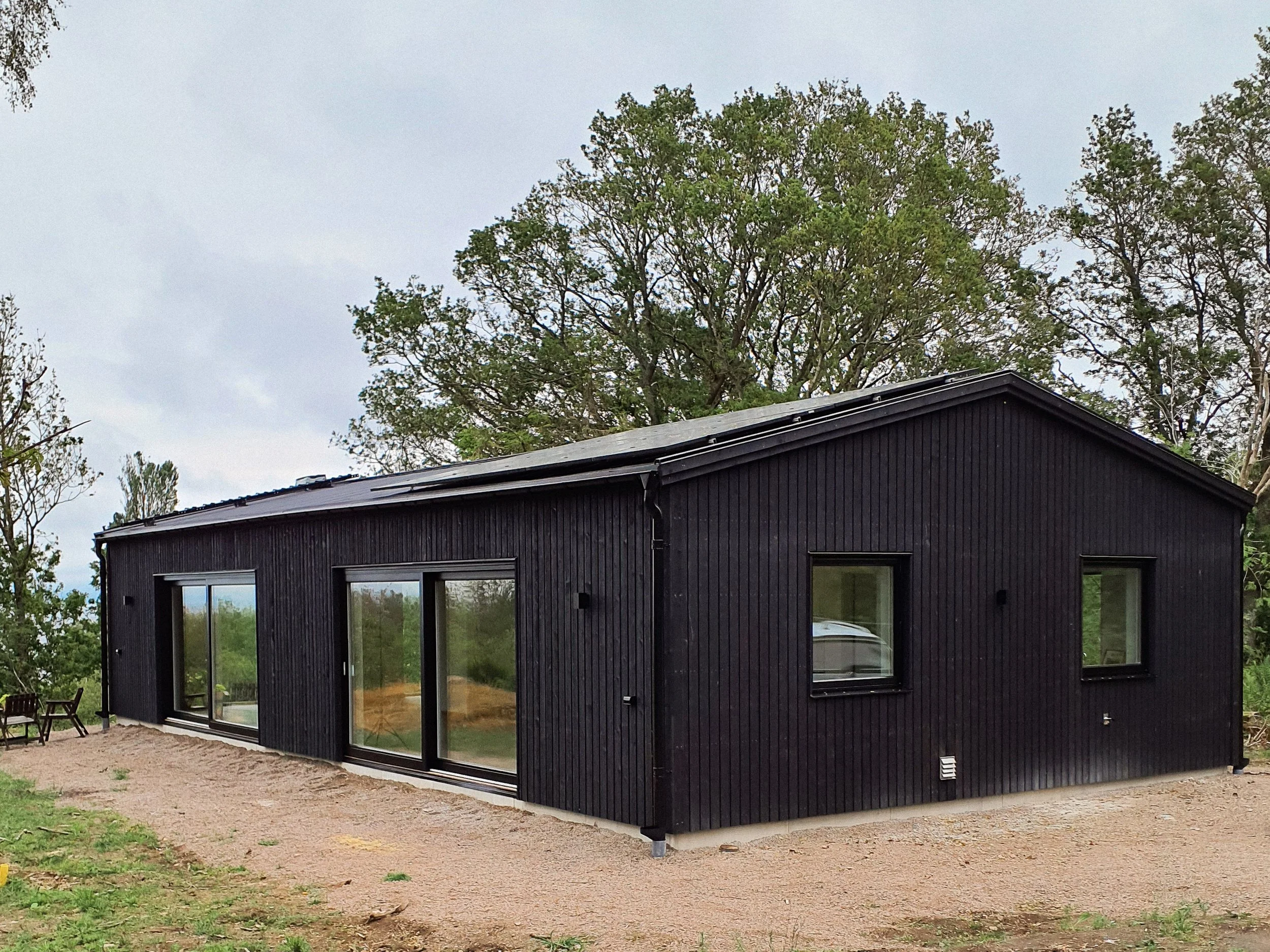 Ett svart hus med stora fönster och en grön natur runt omkring.