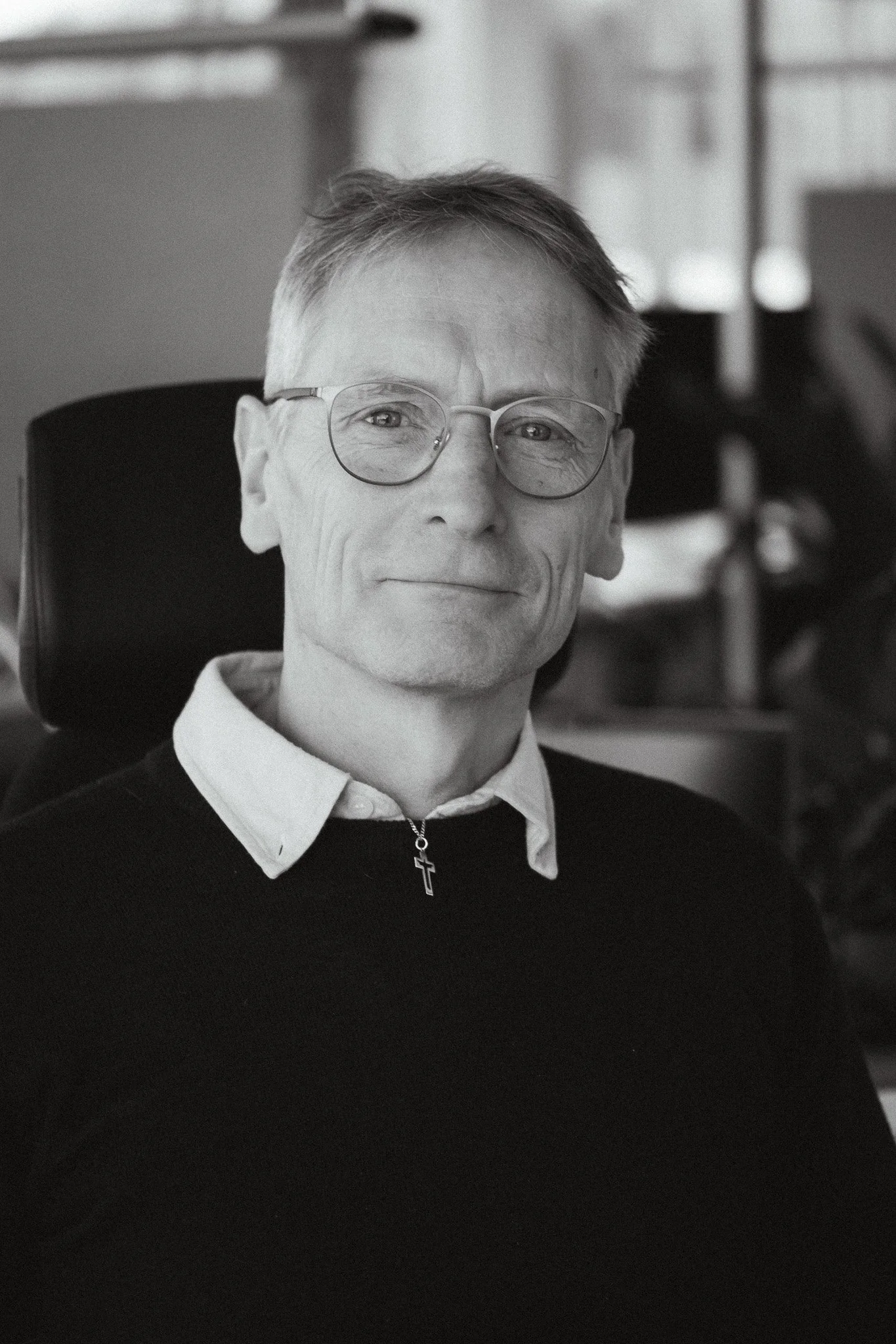 En person med kort ljus hår, glasögon, och en korshalskedja, sittandes i en svart kontorsstol, klädd i en stickad tröja, i ett väl upplyst rum.