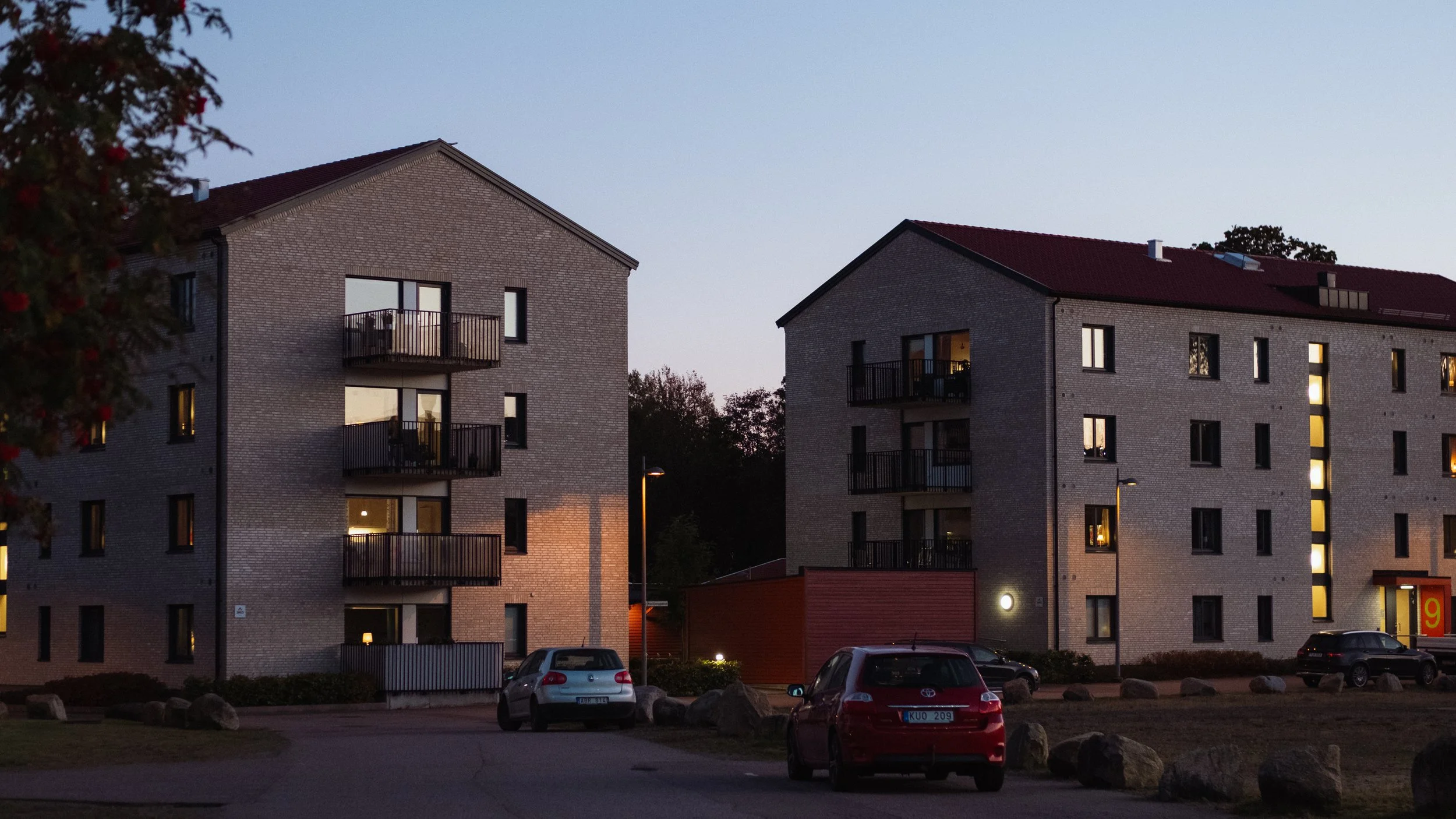 Två trevåningshus av tegel med balkonger, parkerade bilar framför, tidig kväll eller skymning, utomhusbelysning.