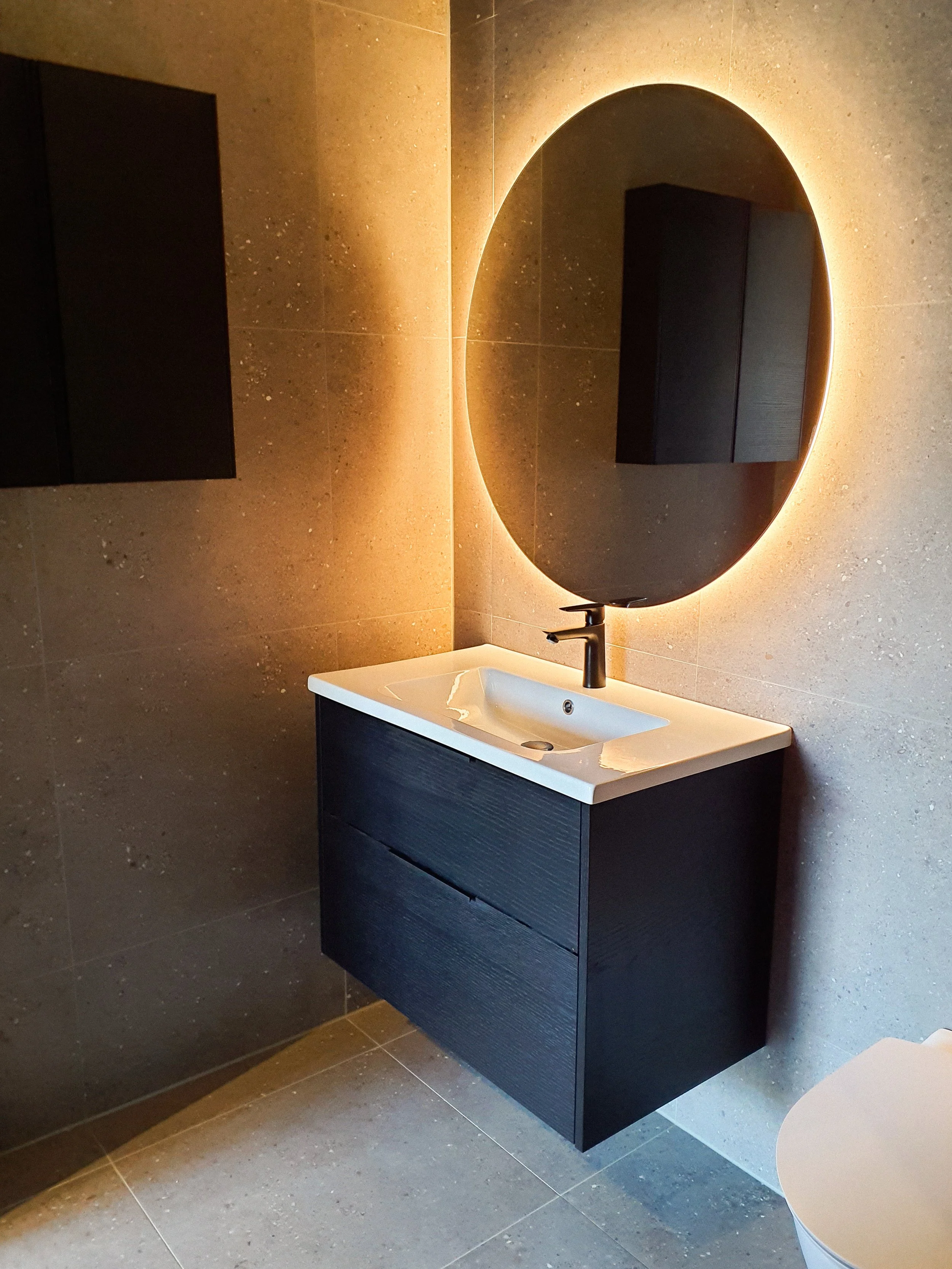 Modern badrum med svart väggskåp, runt spegel med bakgrundsbelysning, beige kakelväggar och golv, vit handfat och svart kran.