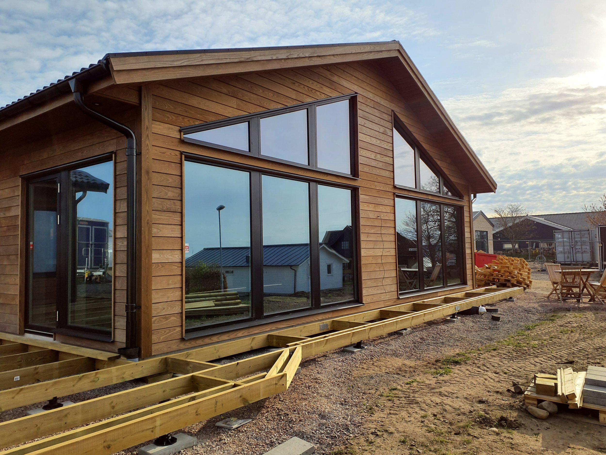 Byggarbetsplats med ett nybyggt hus med stora glasfönster, träfasad och pågående altanunderbyggnad, omgiven av byggmaterial och verktyg.