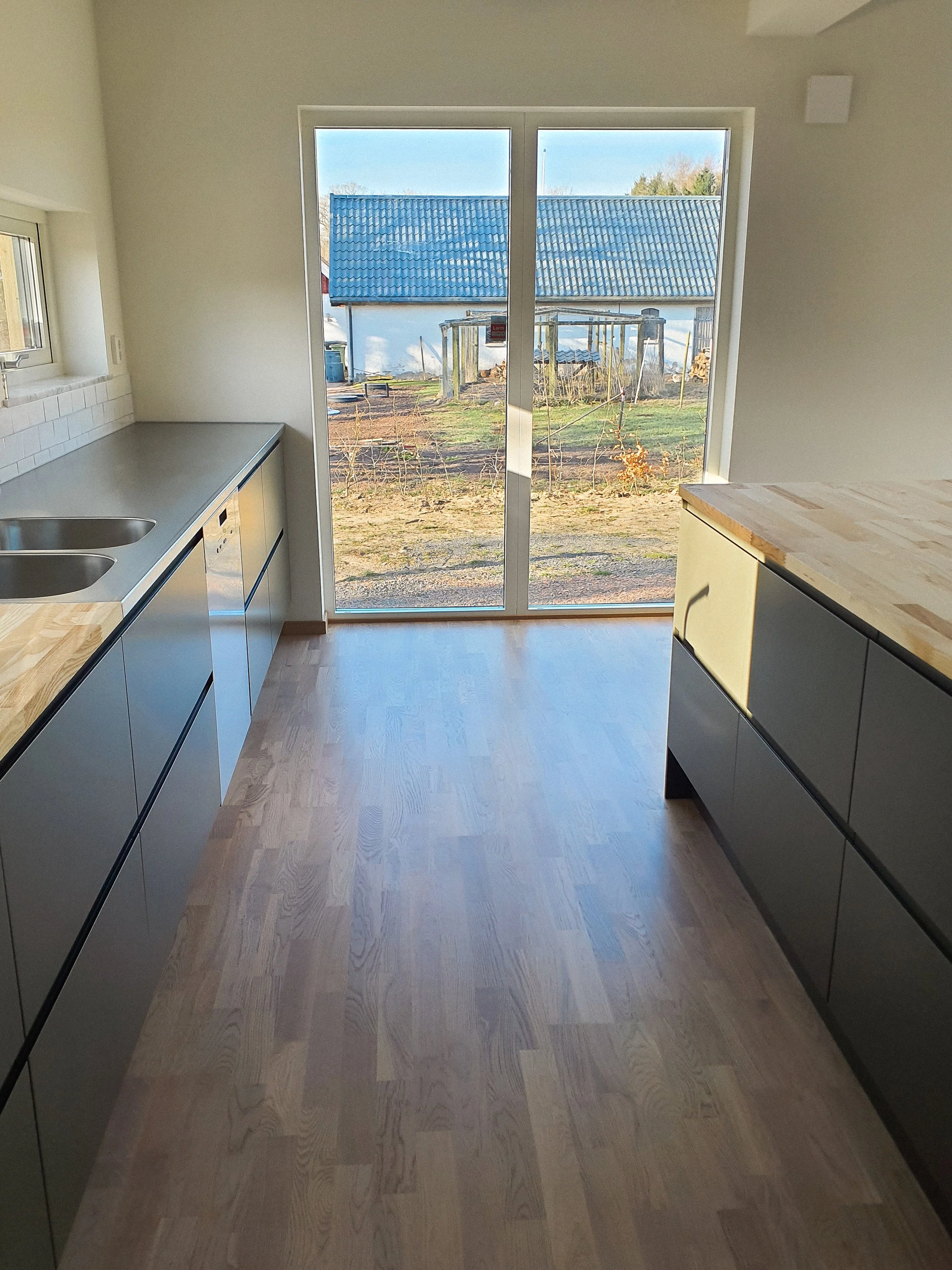 Ett tomt kök med moderna skåp och ljus träbänkar, stort glasdörr mot en trädgård med en liten byggnad i bakgrunden.