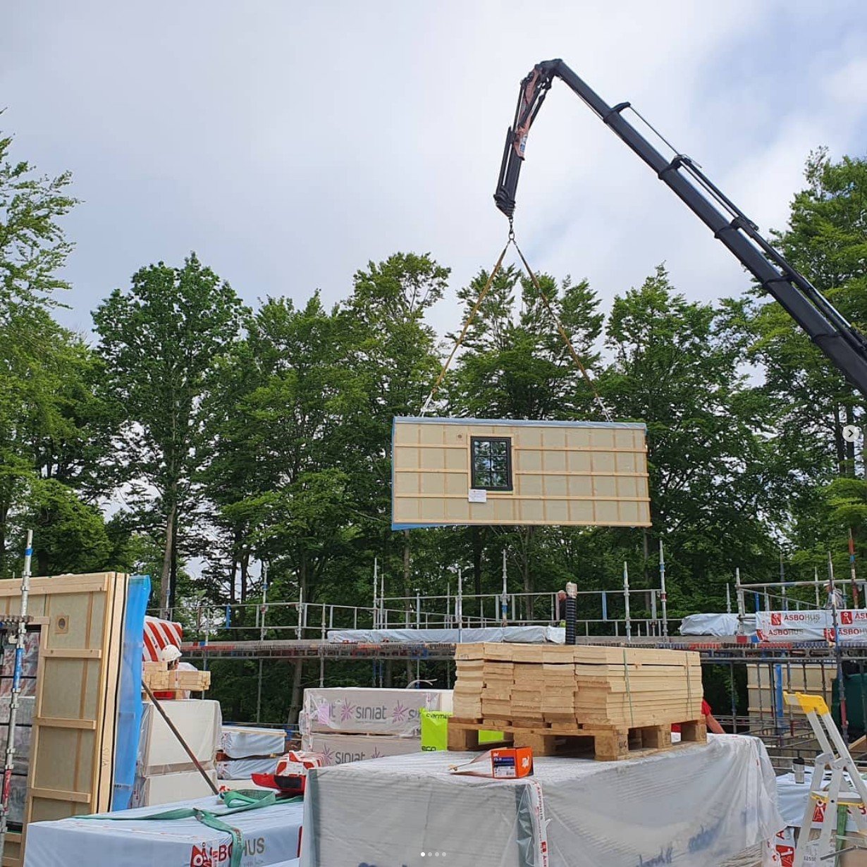 En byggarbetsplats med ett stort gävlar som lyfter en prefab vägg med ett fönster, omringad av byggmaterial och träd i bakgrunden.